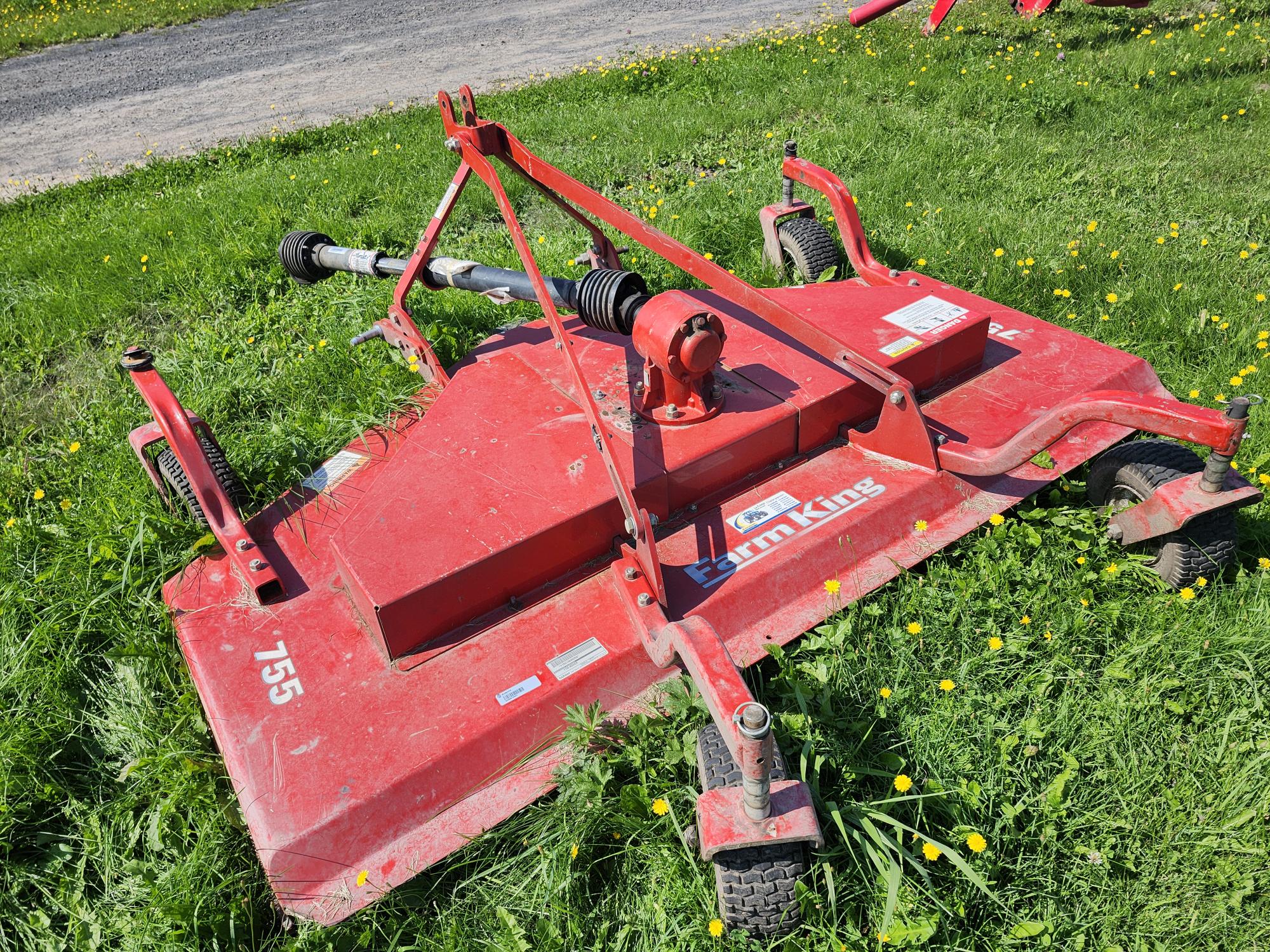 Une tondeuse rotative rouge est posée sur un terrain herbeux. Elle est étiquetée « 755 » et « Yimkins » et possède des roues et un arbre visibles. Des fleurs jaunes parsèment l'herbe verte qui l'entoure.