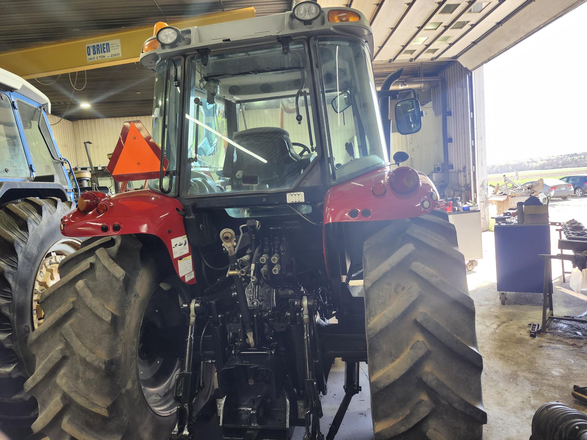 Un tracteur rouge avec de gros pneus arrière est garé dans un garage-atelier. Outils et équipements sont visibles tout autour, et la lumière du soleil entre par une porte ouverte sur la droite.