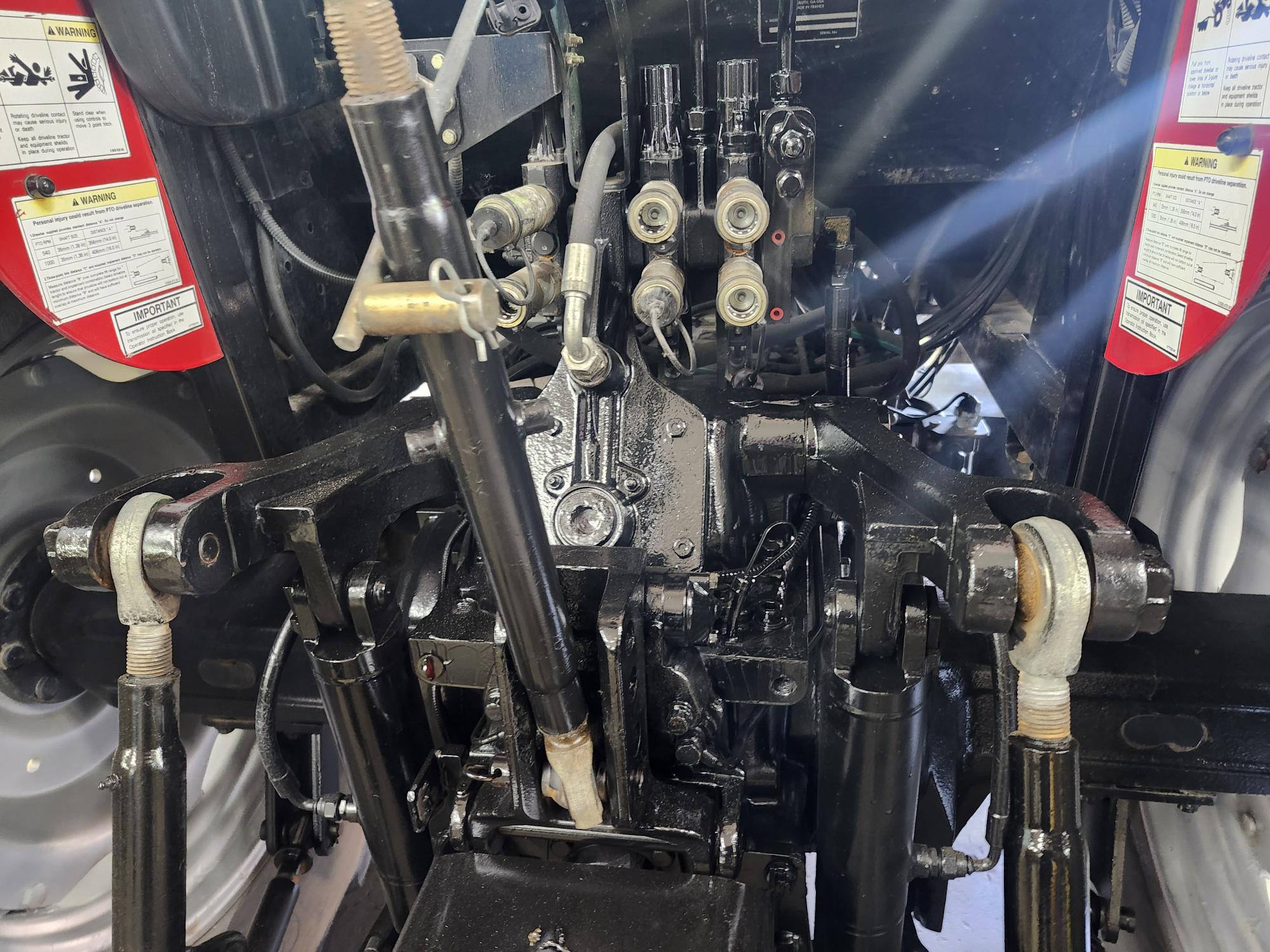 Gros plan du système hydraulique arrière et de l'attelage trois points d'un tracteur, montrant divers leviers de commande, connecteurs hydrauliques et mécanismes, avec des étiquettes d'avertissement visibles des deux côtés.