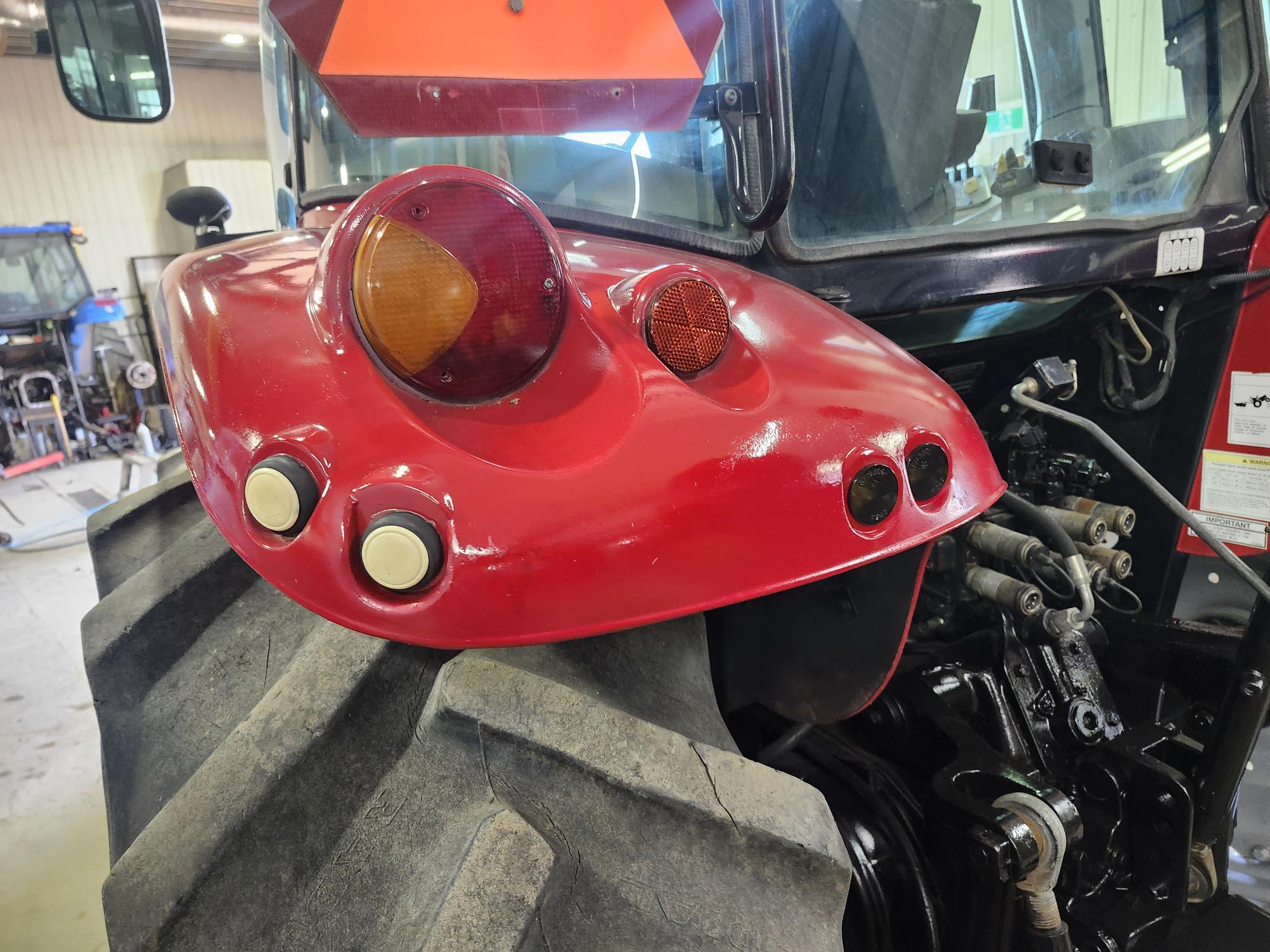 Gros plan du côté arrière gauche d'un tracteur rouge, montrant de gros pneus, un garde-boue rouge avec deux feux circulaires, deux réflecteurs et deux boutons blancs circulaires, avec des machines et un garage en arrière-plan.