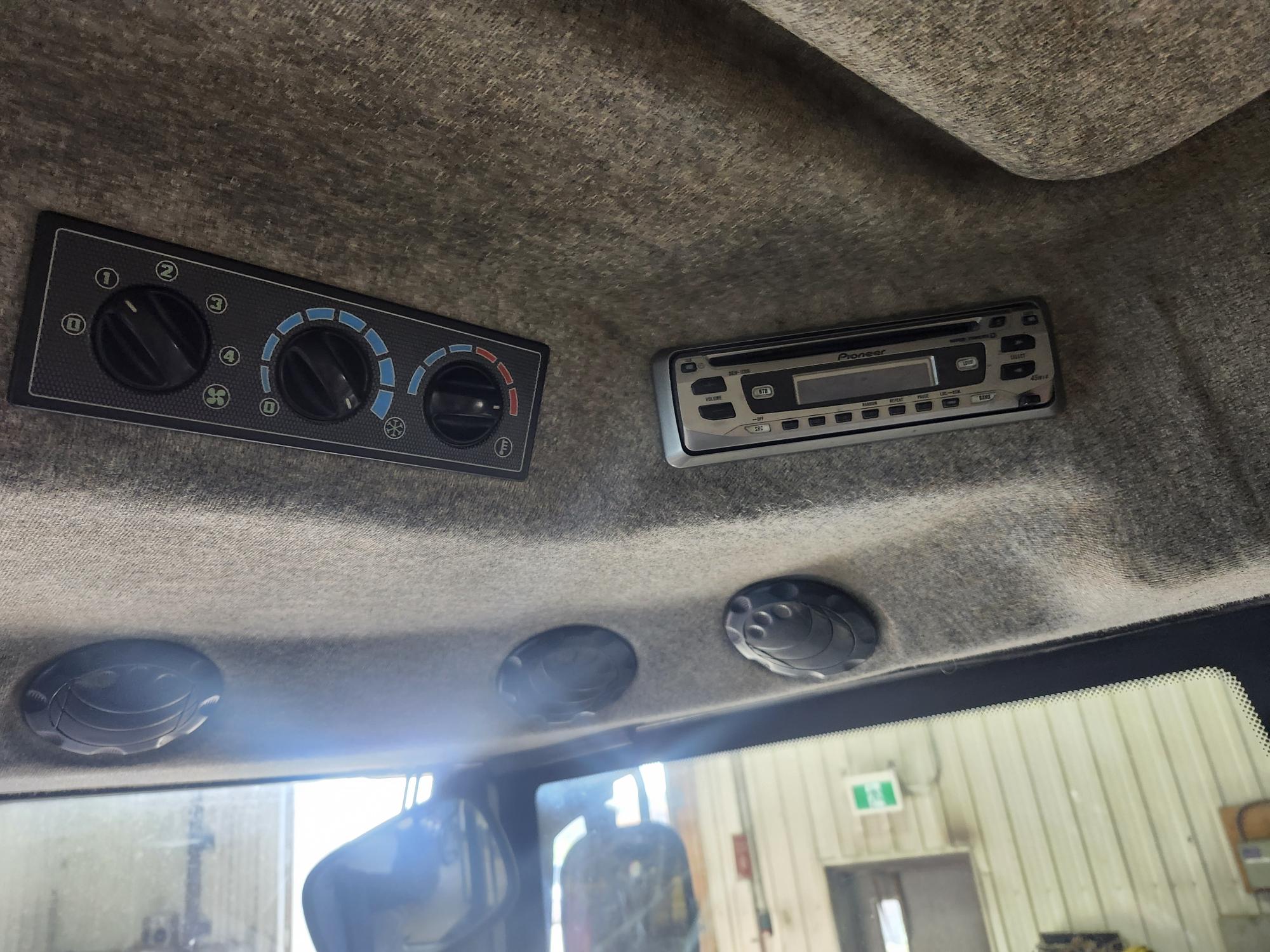 Deux panneaux de commande avec cadrans et boutons, dont un pour la température et une radio, sont fixés au plafond recouvert de tissu à l'intérieur d'un véhicule. Trois bouches d'aération circulaires et une fenêtre donnant sur un garage intérieur sont également visibles.