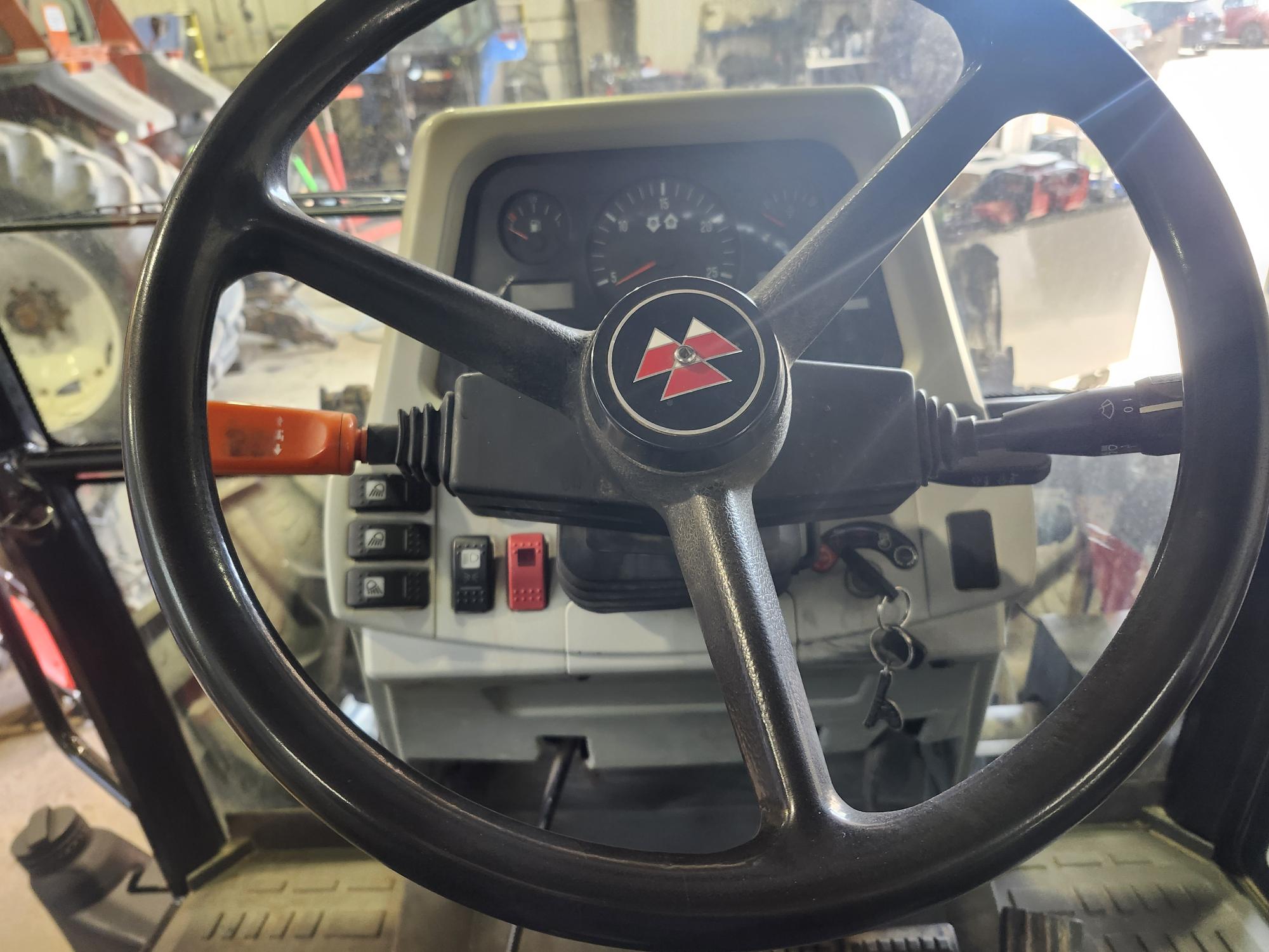 Vue rapprochée du volant et du tableau de bord d'un tracteur Massey Ferguson, montrant divers boutons de commande, jauges et la clé de contact insérée, avec l'équipement d'atelier visible en arrière-plan.