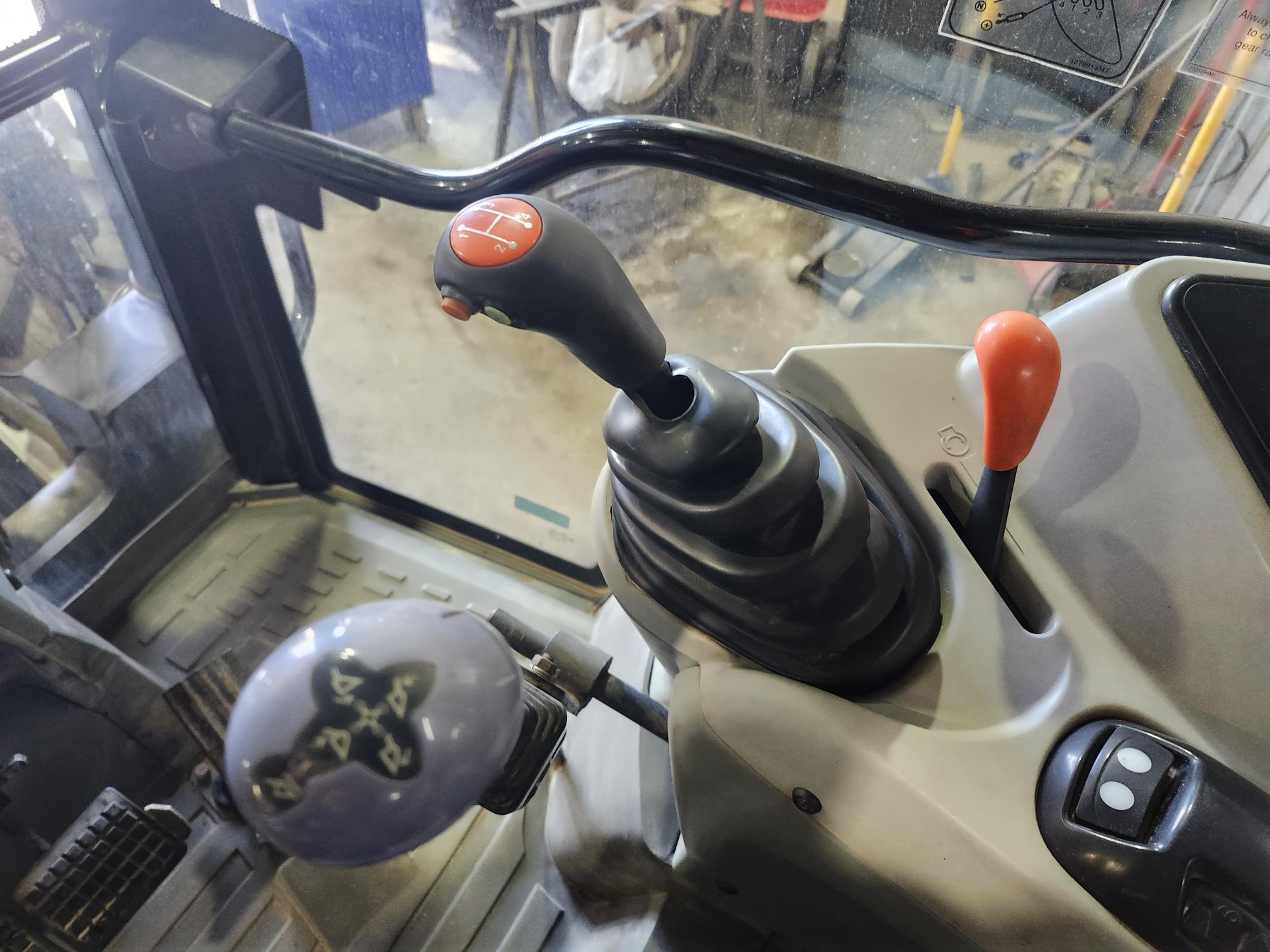 Gros plan de l'intérieur d'un tracteur montrant le levier de vitesses, le joystick et les leviers de commande, avec les pédales au sol et l'équipement de l'atelier visible à travers la fenêtre de la cabine en arrière-plan.