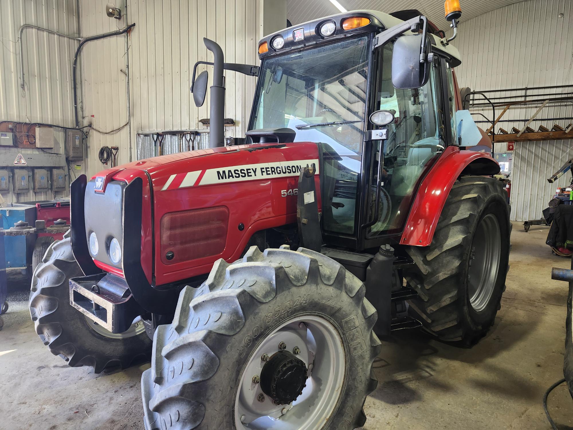 Un tracteur rouge Massey Ferguson 5465 est garé à l'intérieur d'un garage ou d'un atelier avec des outils et de l'équipement visibles en arrière-plan.