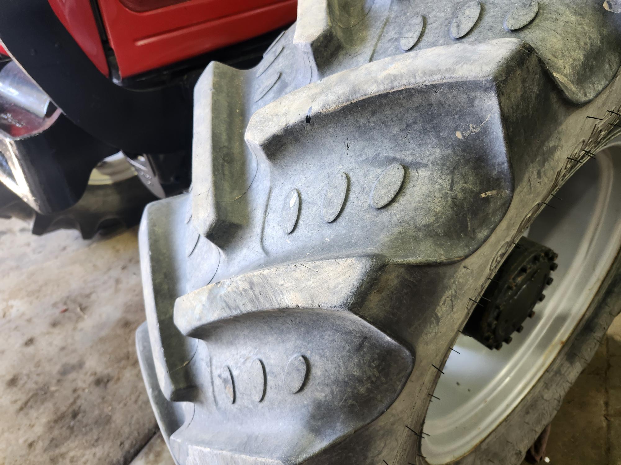 Gros plan d'un gros pneu de tracteur robuste aux sculptures profondes, fixé à un tracteur rouge. Le pneu est légèrement poussiéreux et le véhicule est garé sur un sol en béton à l'intérieur.