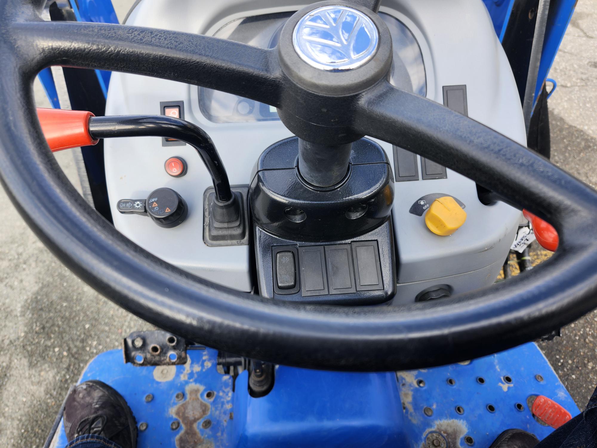 Gros plan sur le tableau de bord d'un tracteur, avec un grand volant avec un emblème bleu. Diverses commandes et boutons sont visibles, notamment un levier noir avec une poignée rouge et un bouton jaune. Le sol est partiellement visible en dessous.