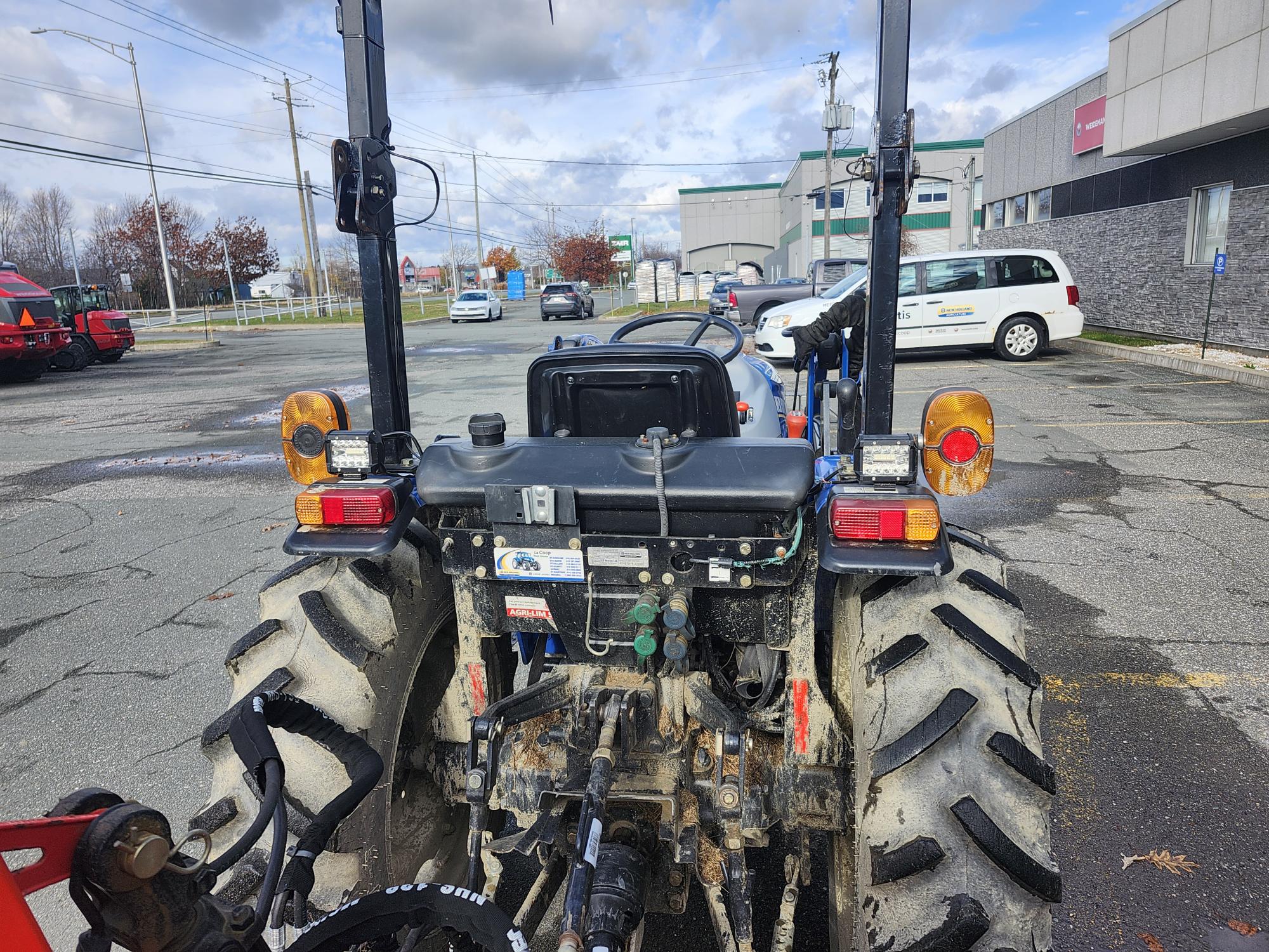 L'image montre l'arrière d'un tracteur stationné dans un parking, avec des lumières et des leviers de commande visibles. L'arrière-plan comprend plusieurs véhicules stationnés et un bâtiment commercial sous un ciel partiellement nuageux.