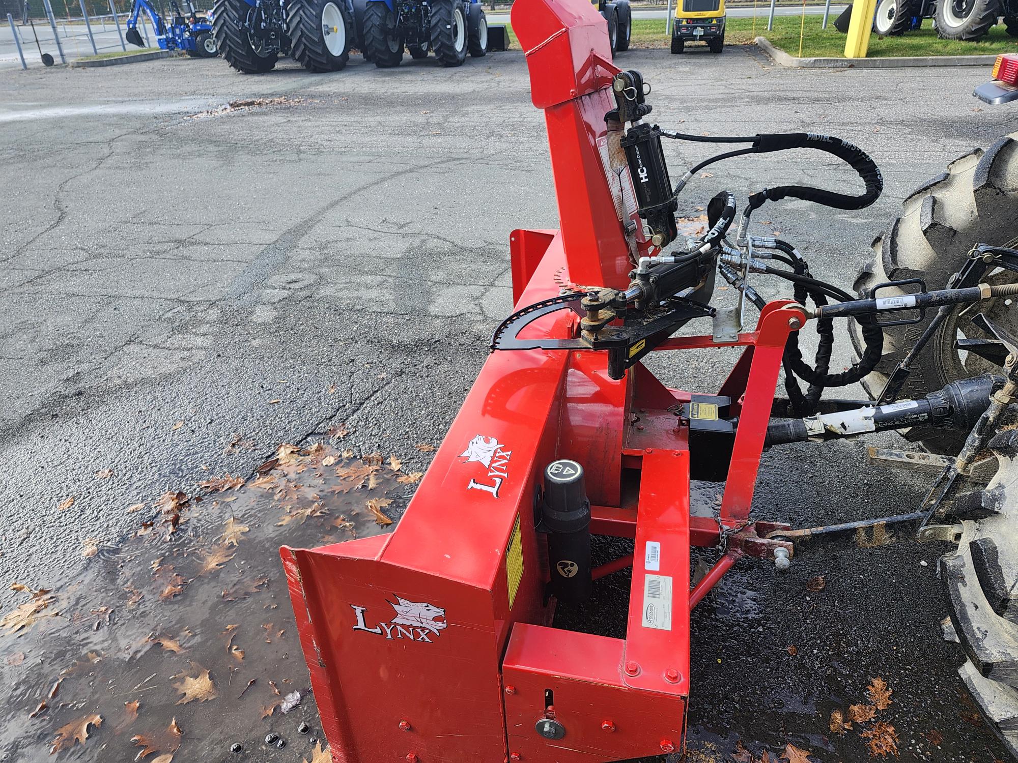 Une souffleuse à neige Lynx rouge attachée à un tracteur. Elle est garée sur une surface asphaltée mouillée avec des feuilles éparpillées. À l'arrière-plan, il y a plusieurs gros pneus et une zone clôturée.