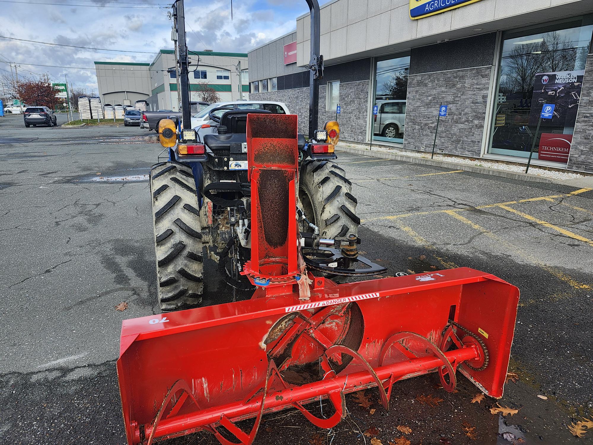 Une souffleuse à neige rouge est attachée à l'arrière d'un tracteur dans un parking vide. Le tracteur est garé près d'un bâtiment avec plusieurs devantures de magasins et un ciel nuageux au-dessus.