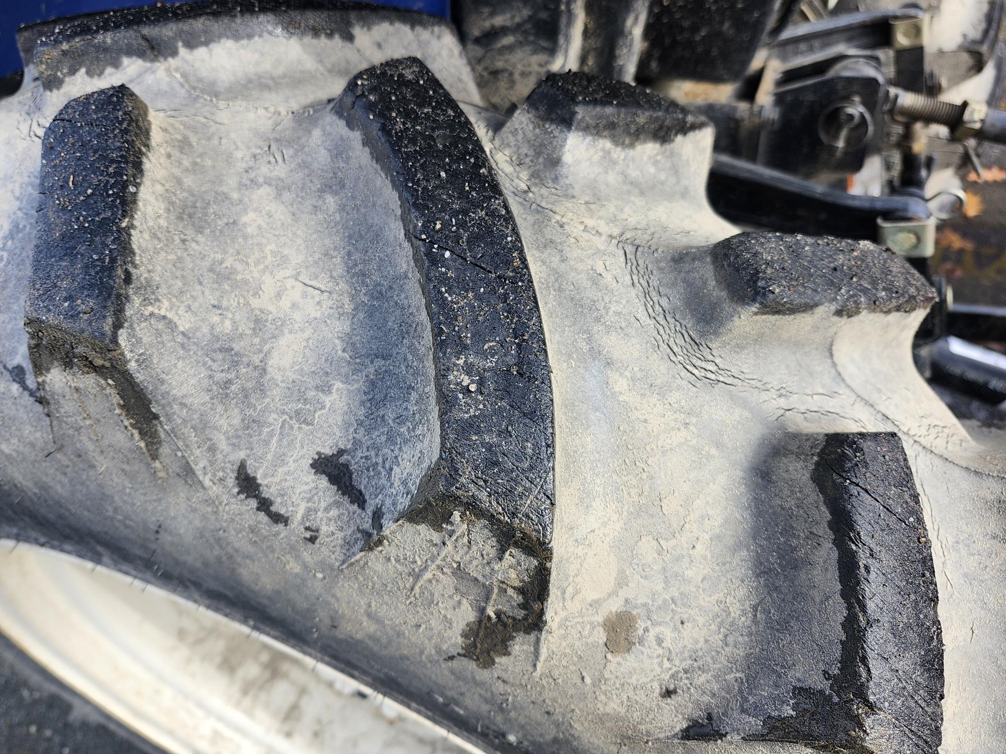 Gros plan d'un pneu de tracteur usé et boueux avec des bandes de roulement distinctes. Les grandes rainures sont visibles, soulignant la texture rugueuse et la saleté incrustée dans la surface. L'arrière-plan comprend des traces de pièces de machines.