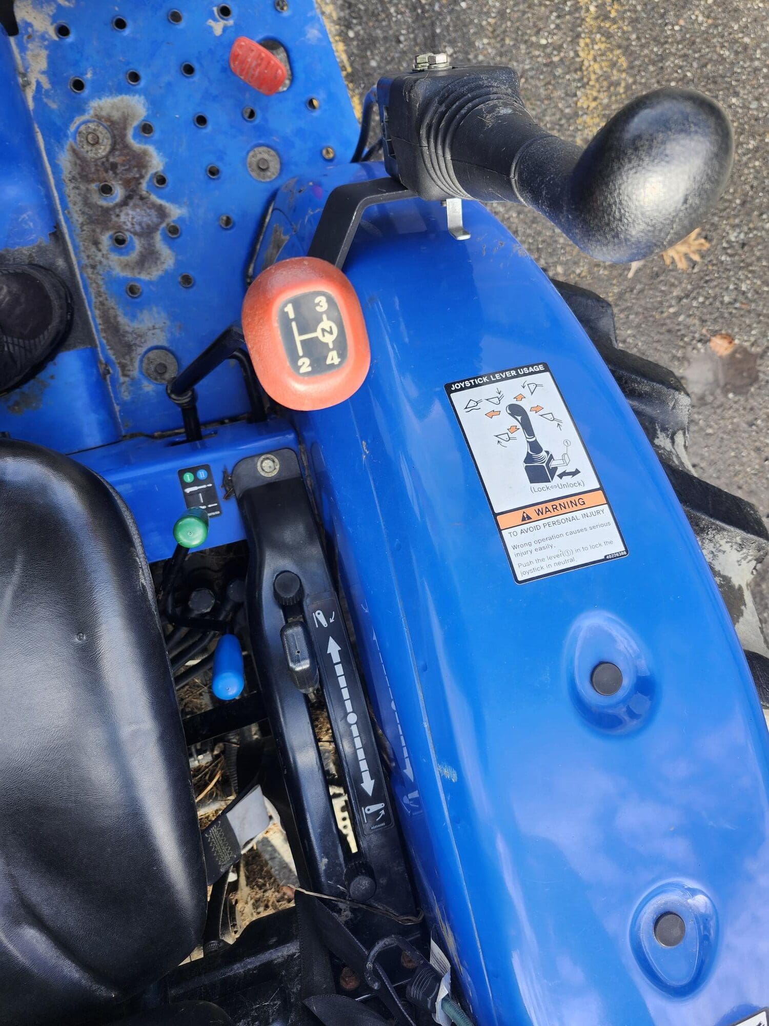 Vue rapprochée du siège et des commandes d'un tracteur bleu, avec un levier de vitesse avec des positions étiquetées. Un autocollant d'avertissement avec des schémas est visible, ainsi qu'un repose-pied en métal texturé et une colonne de direction.