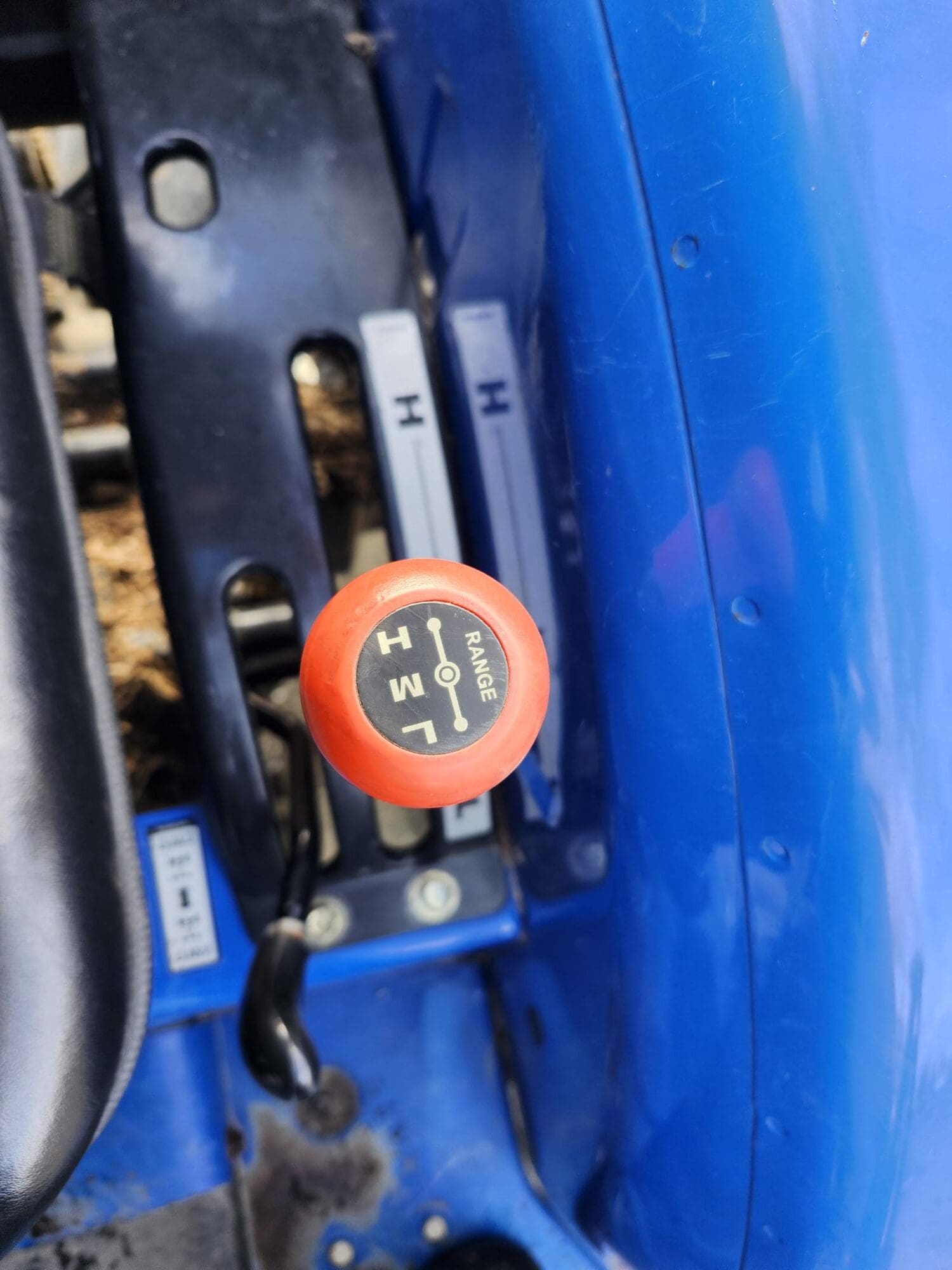 Gros plan d'un levier de vitesse sur une machine bleue, avec un bouton rouge étiqueté avec les différents réglages de plage : M (milieu), H (élevé) et L (bas). L'arrière-plan montre une partie de la surface métallique de la machine.