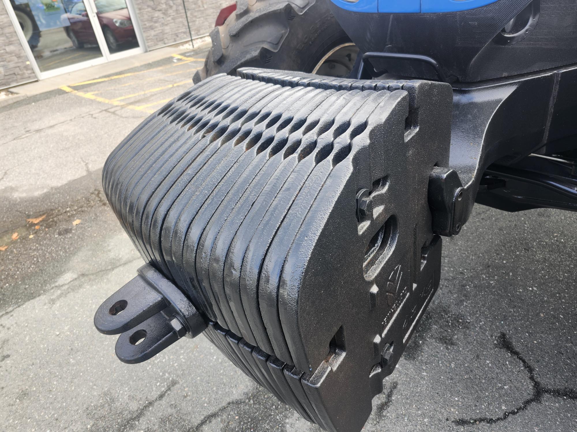 Gros plan sur des plaques métalliques empilées fixées à l'avant d'un tracteur bleu, servant de contrepoids. Les plaques sont conçues de manière uniforme avec des rainures et des fentes visibles, positionnées sur un parking avec un bâtiment en arrière-plan.