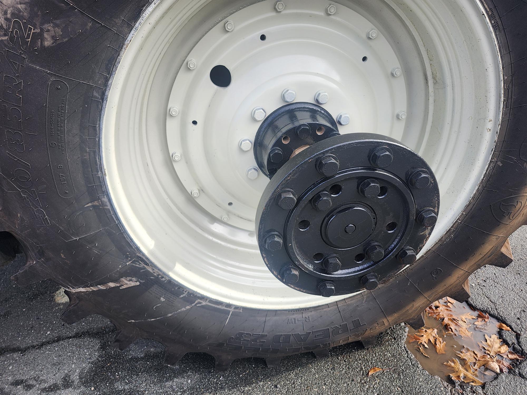 Gros plan d'un gros pneu de tracteur avec un moyeu de roue noir, montrant des motifs de bande de roulement détaillés. Le pneu est sur une surface asphaltée mouillée avec des feuilles brunes tombées à proximité.