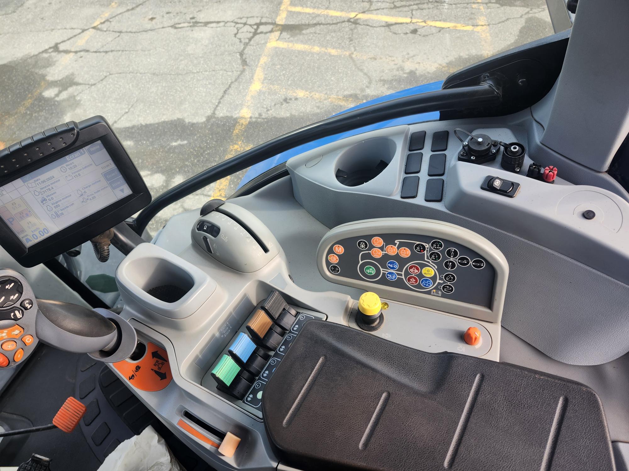 Vue intérieure d'une cabine de tracteur moderne montrant un tableau de bord avec diverses commandes et boutons colorés, un volant, un écran tactile et un siège confortable. Le sol et une partie du pare-brise sont également visibles.