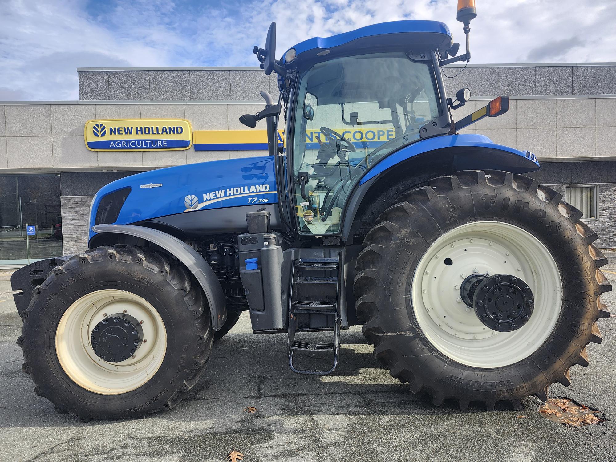 Un tracteur New Holland bleu équipé de gros pneus est garé devant une concession agricole New Holland. Les vitres du tracteur sont réfléchissantes et le bâtiment de la concession est visible en arrière-plan.