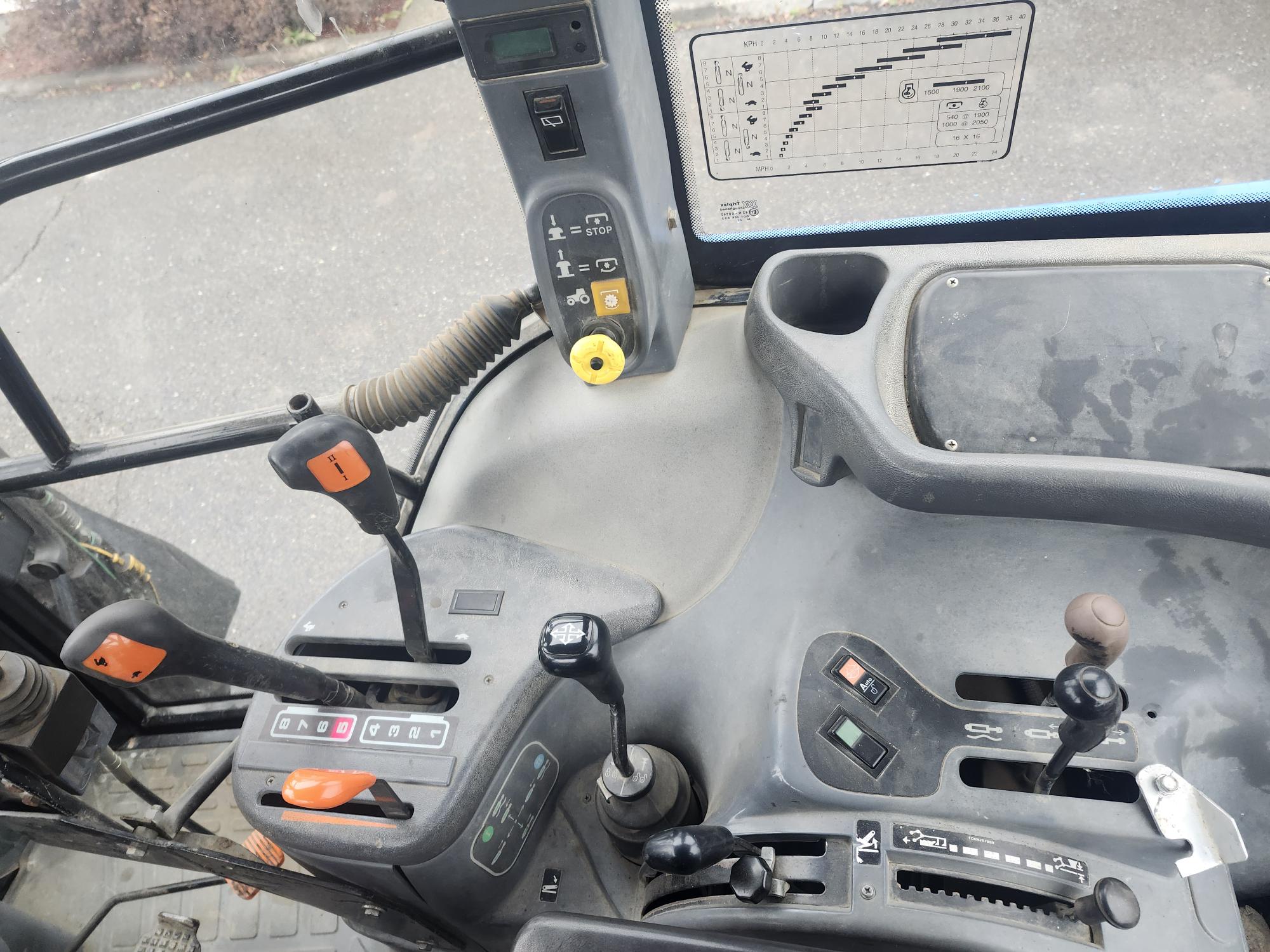 L'image montre le panneau de commande intérieur d'un véhicule, peut-être un tracteur ou une machine lourde. Différents leviers, boutons et interrupteurs sont visibles sur un tableau de bord, ainsi qu'un levier de vitesse étiqueté avec des lettres et des chiffres.