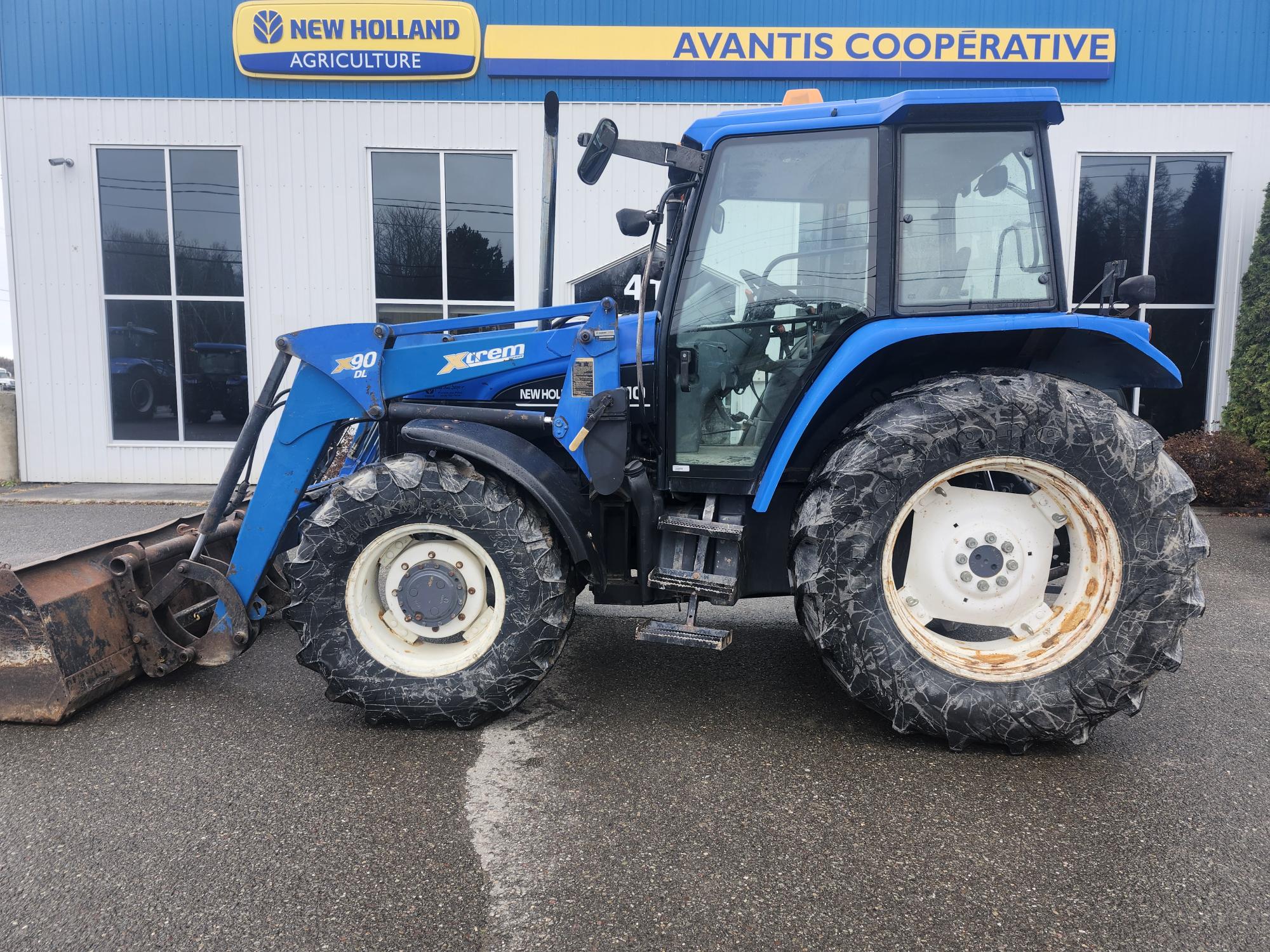 Un tracteur New Holland bleu avec de gros pneus est garé sur un trottoir mouillé devant un bâtiment sur lequel sont inscrits « New Holland Agriculture » et « Avantis Cooperative ». Le tracteur est équipé d'un chargeur frontal avec un godet rouillé.