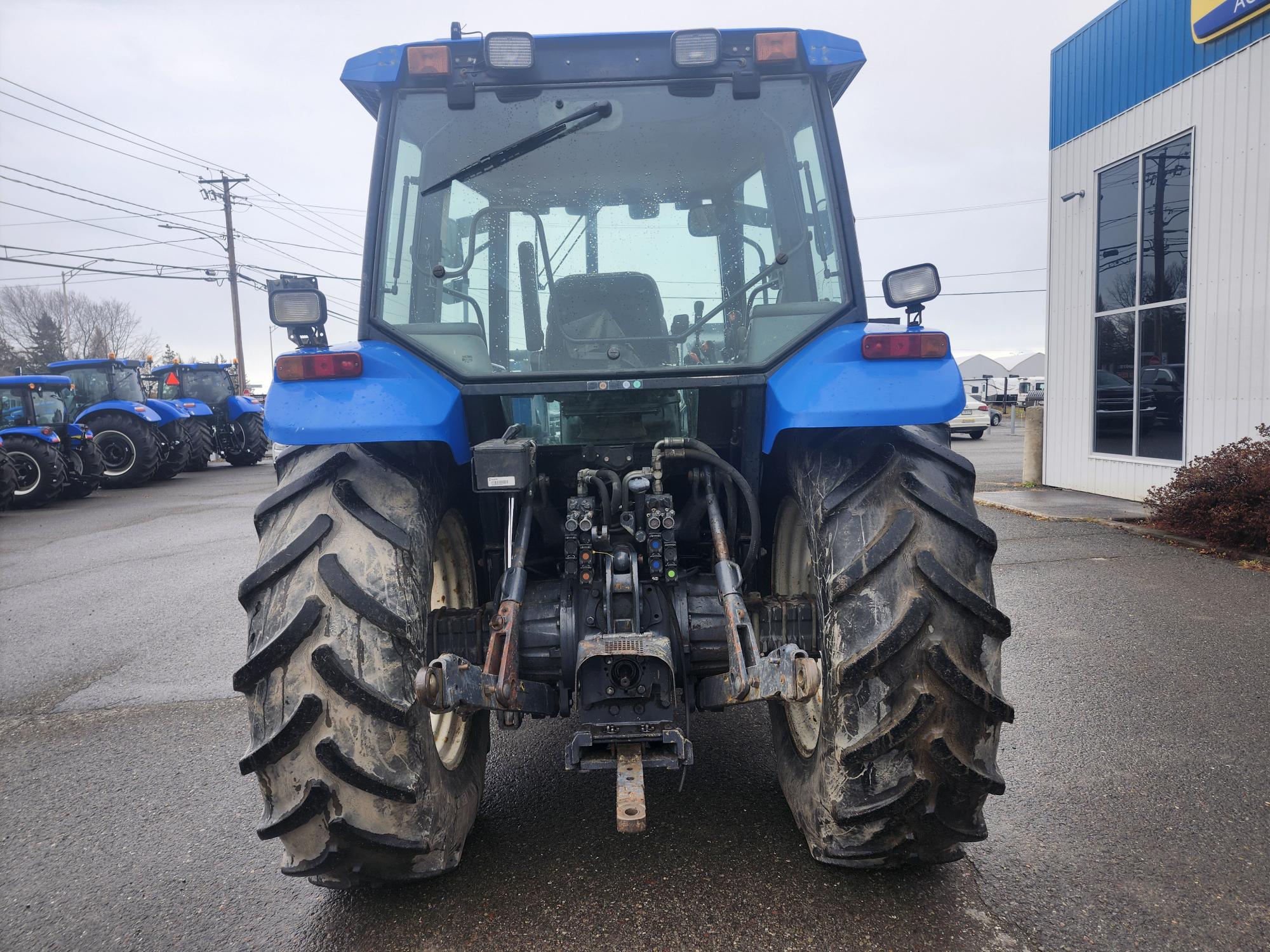 Vue arrière d'un tracteur bleu avec de gros pneus, stationné sur une chaussée mouillée près d'un concessionnaire. Le tracteur est équipé de divers composants de machines et d'autres tracteurs sont visibles en arrière-plan.