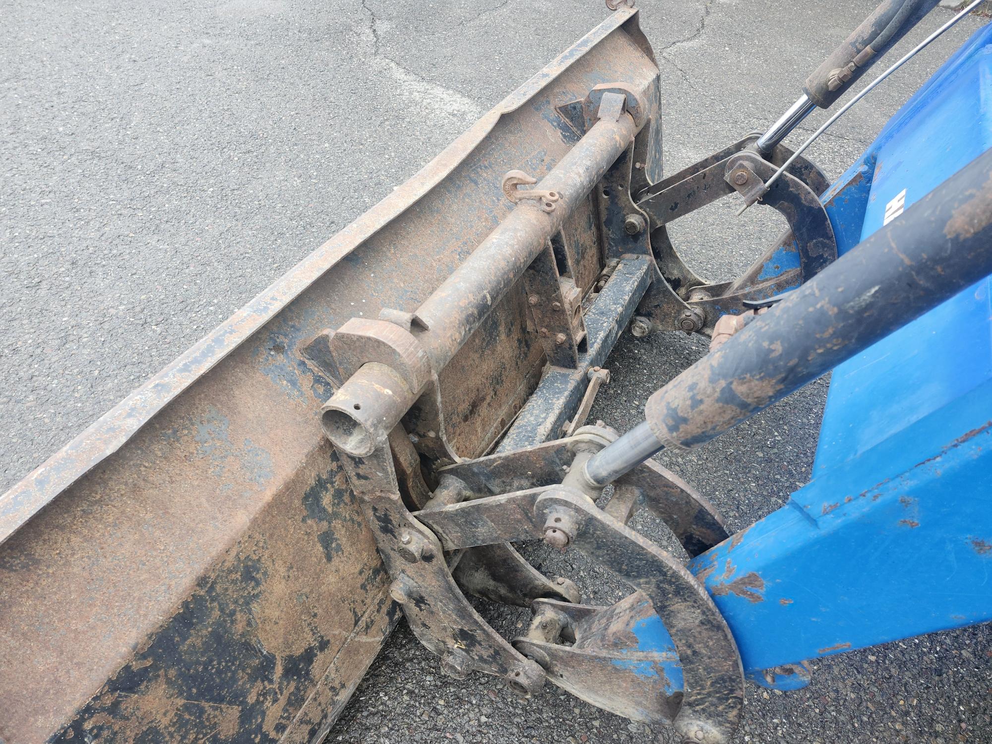 Gros plan d'un chasse-neige rouillé, bleu et marron sur une surface en béton, montrant des tuyaux hydrauliques et des joints métalliques.