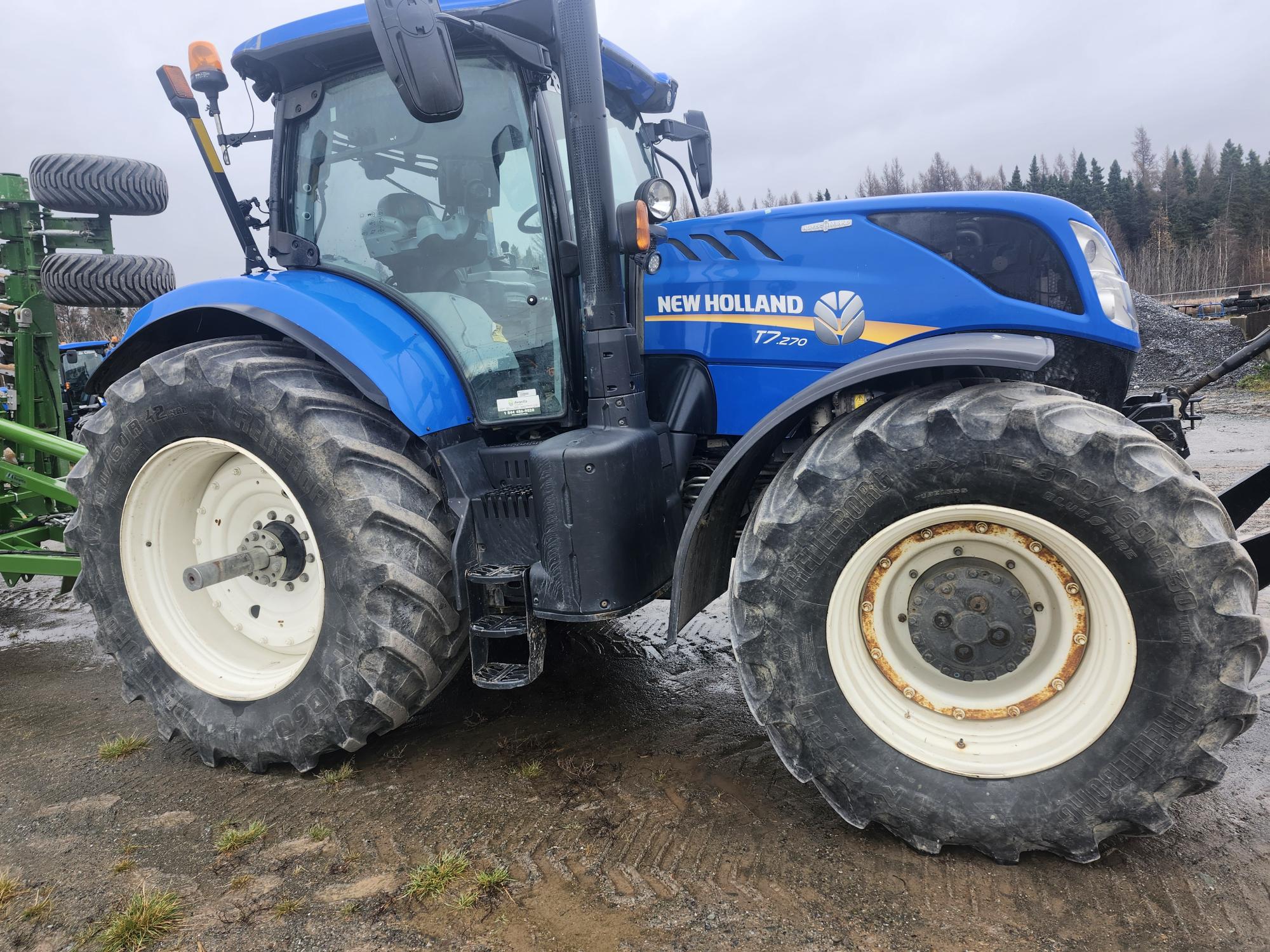 Un tracteur New Holland T7.270 bleu est stationné sur une surface boueuse. Le tracteur est équipé de gros pneus et est positionné près d'un terrain boisé avec un ciel couvert.