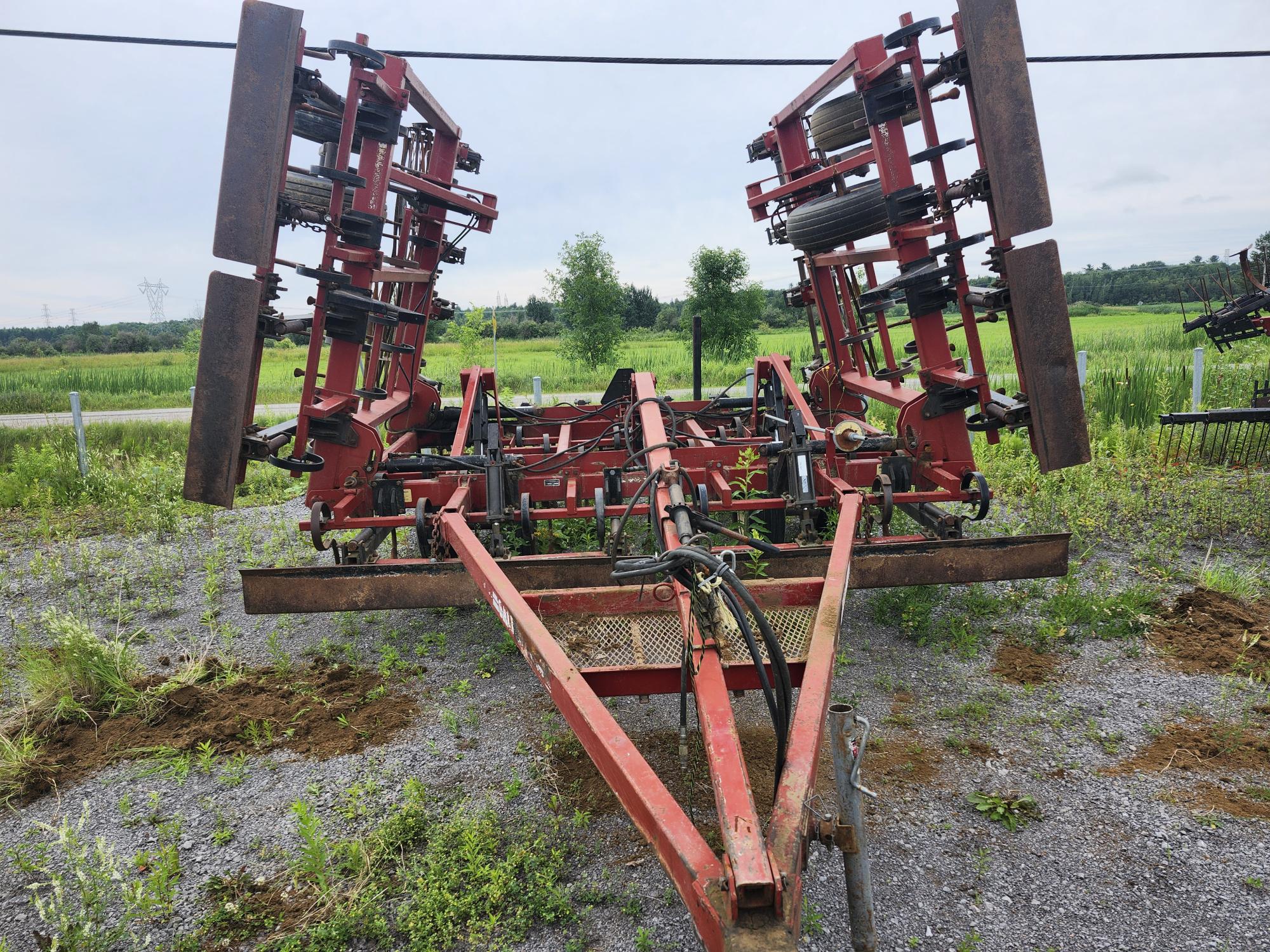 Machines agricoles rouges sur une surface de gravier, avec des composants métalliques verticaux et des systèmes hydrauliques, entourées de verdure et d'un ciel clair.