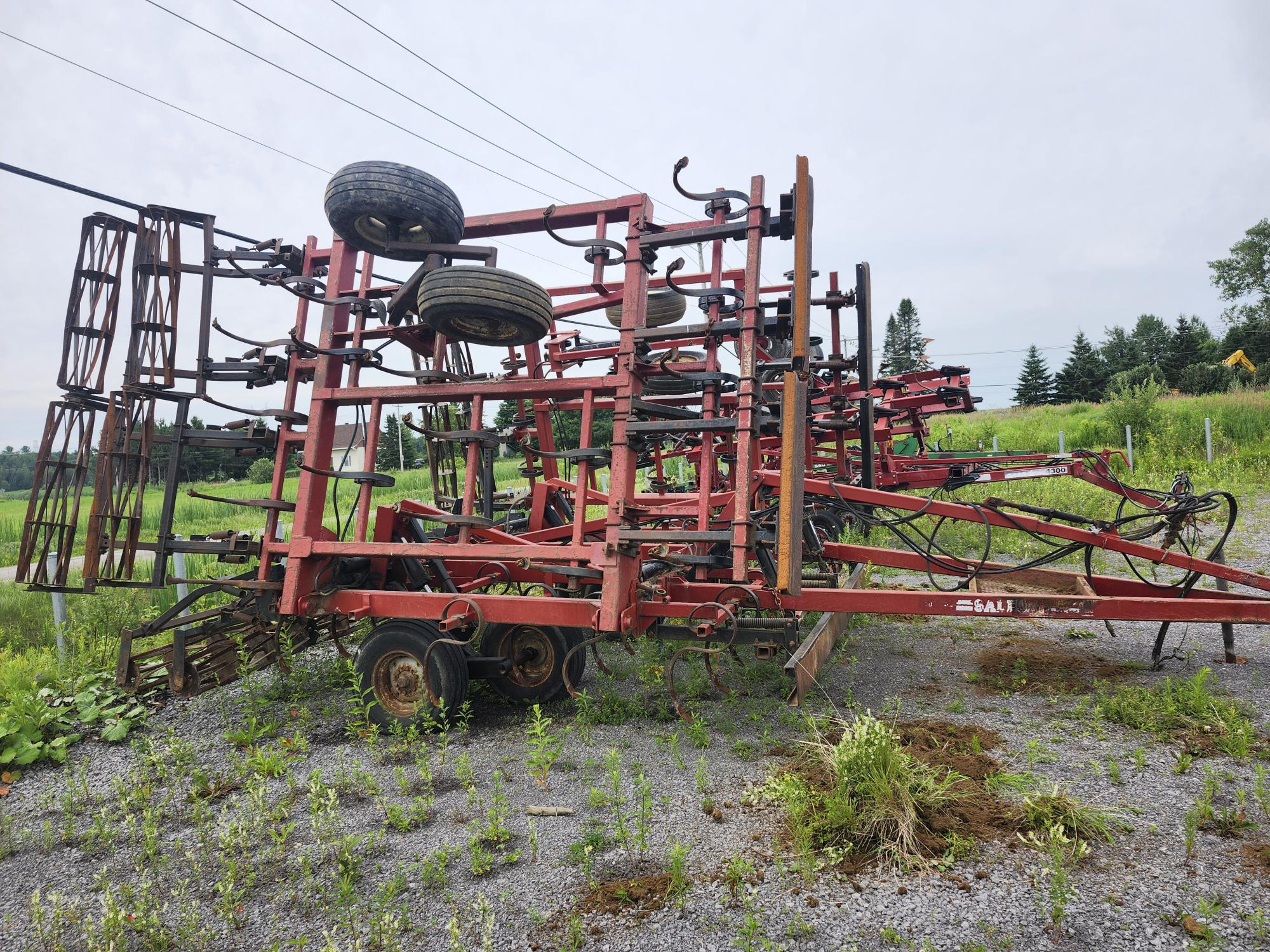 Un gros engin agricole rouge avec plusieurs roues et barres métalliques, garé sur du gravier. Entouré d'herbe verte et d'arbres, avec des lignes électriques visibles en arrière-plan sous un ciel nuageux.