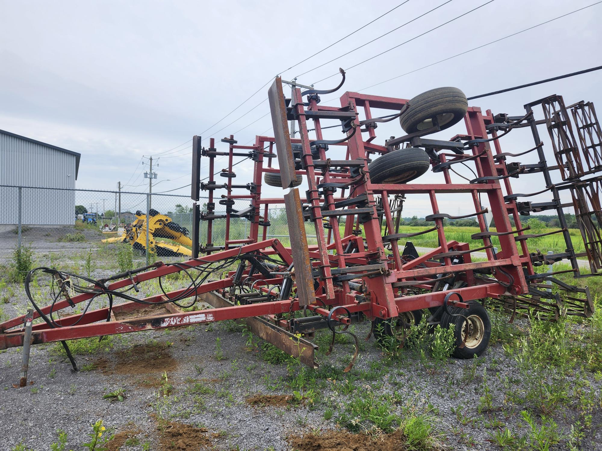 Un outil agricole rouge équipé de pneus, peut-être un cultivateur, repose inutilisé sur une surface en gravier près d'une clôture grillagée. Un bâtiment est visible en arrière-plan, ainsi que d'autres équipements et des parcelles d'herbe.