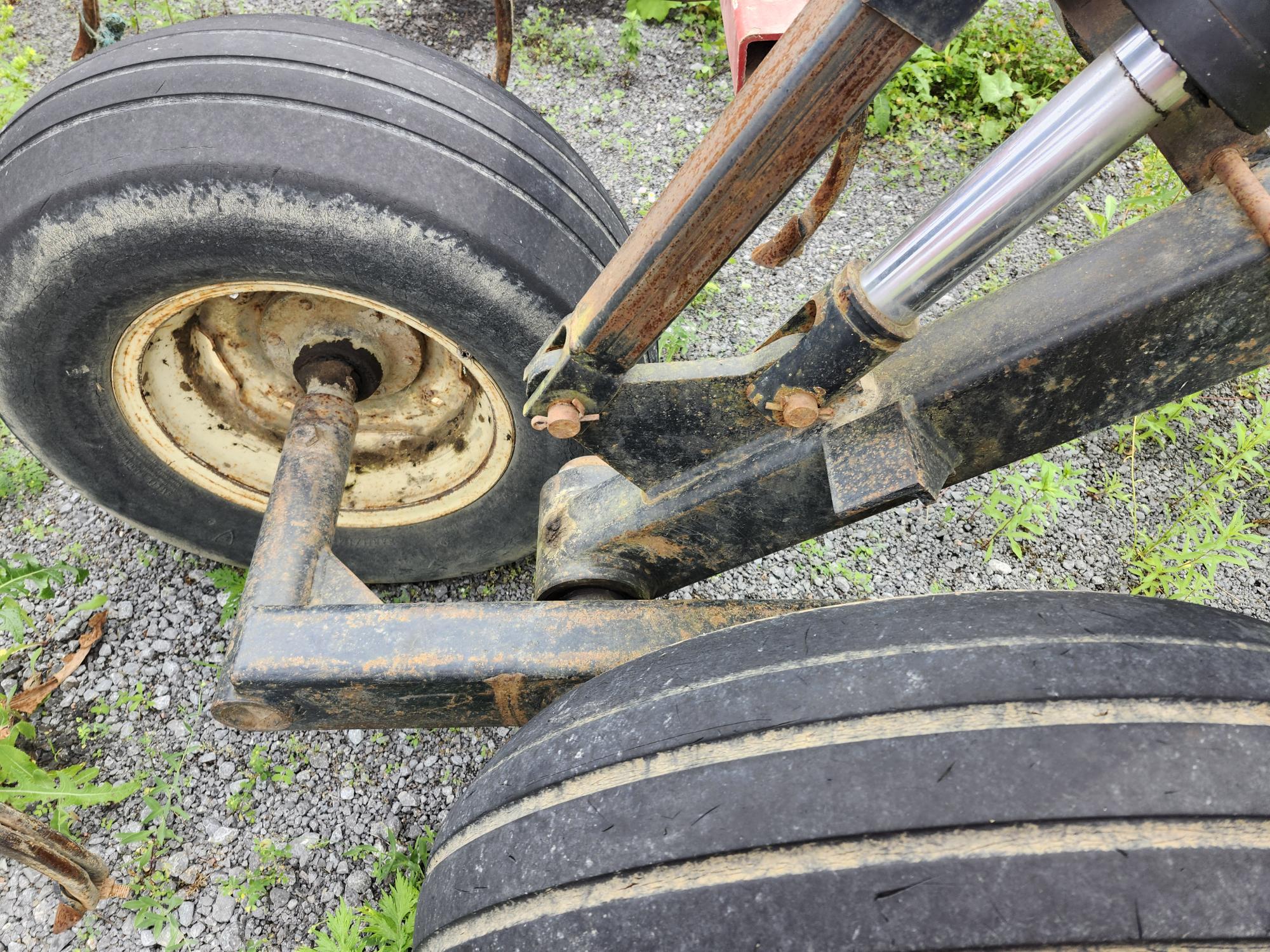 Gros plan sur les roues et le système de suspension d'un véhicule lourd sur du gravier. Les pneus sont usés et les pièces métalliques présentent des signes de rouille et d'usure. De la verdure est visible autour des pierres au sol.