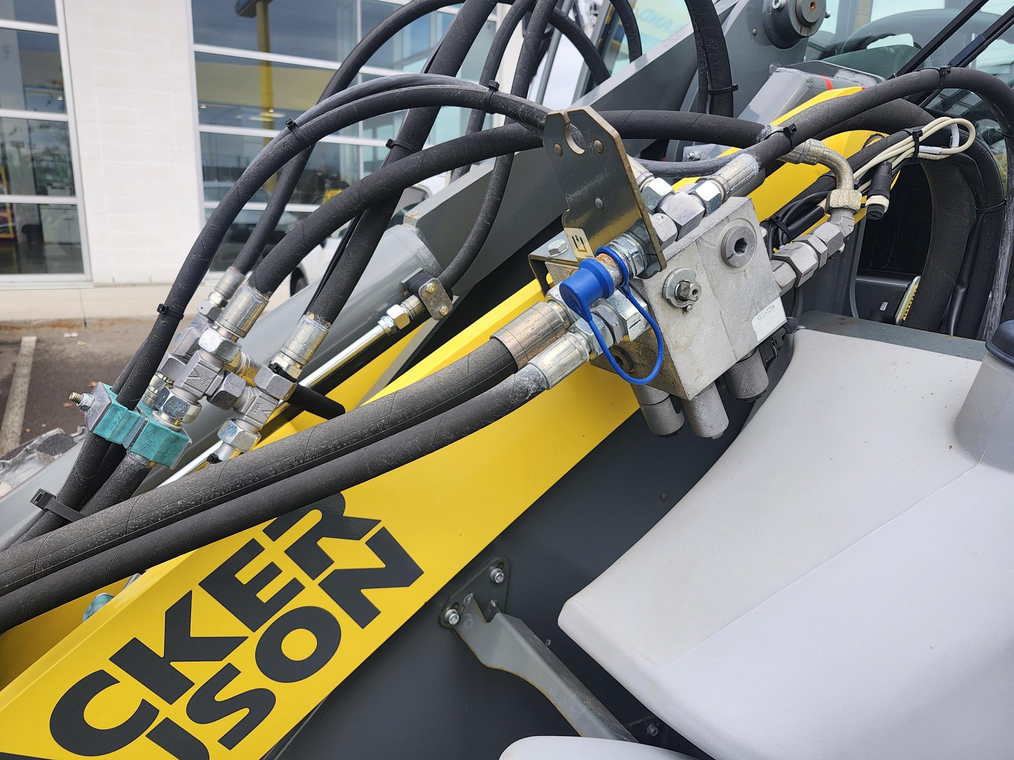 Gros plan sur des tuyaux hydrauliques et des raccords métalliques sur un véhicule de construction jaune, montrant partiellement le nom de la marque « WACKER NEUSON », avec un bâtiment et des fenêtres en arrière-plan.