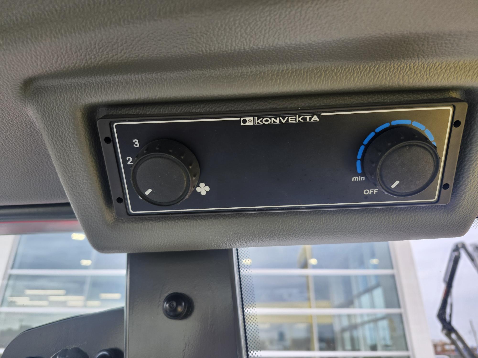 Gros plan d'un panneau de commande Konvekta monté sur le plafond d'un véhicule, comportant deux cadrans noirs pour le réglage de la vitesse du ventilateur et de la température, avec des fenêtres et une partie de l'équipement de construction visibles à l'extérieur.