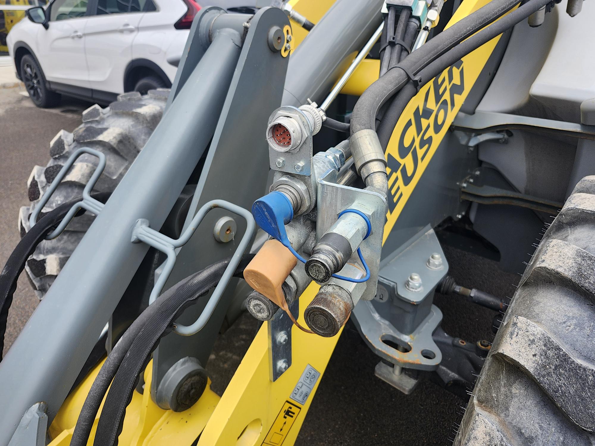 Gros plan des tuyaux hydrauliques et des connecteurs sur le bras d'un véhicule de construction jaune Wacker Neuson, avec une partie d'une voiture blanche visible en arrière-plan.
