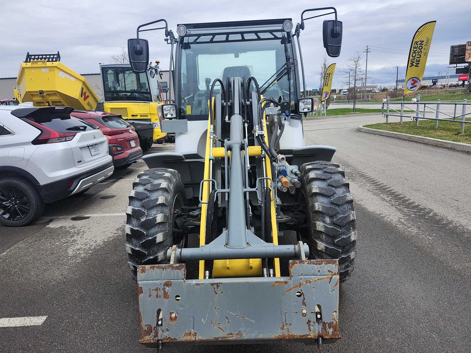 Vue de face d'une grande chargeuse de chantier jaune et grise garée sur le trottoir, avec des voitures et un autre véhicule de chantier à proximité, et un ciel nuageux en arrière-plan.