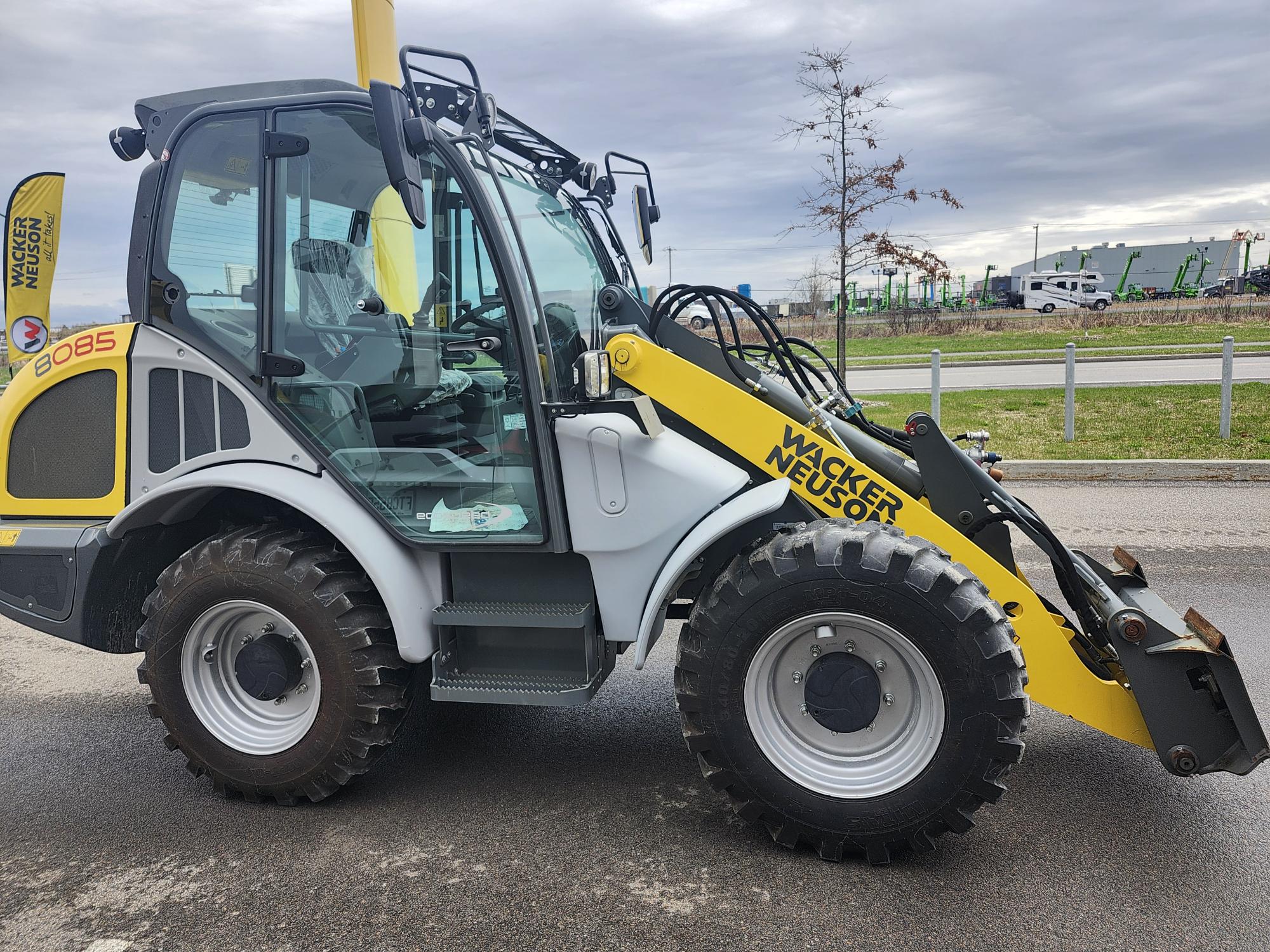 Une chargeuse de chantier Weidemann jaune et grise avec de gros pneus est garée sur une surface pavée près d'une route, avec des équipements industriels et un ciel nuageux en arrière-plan.