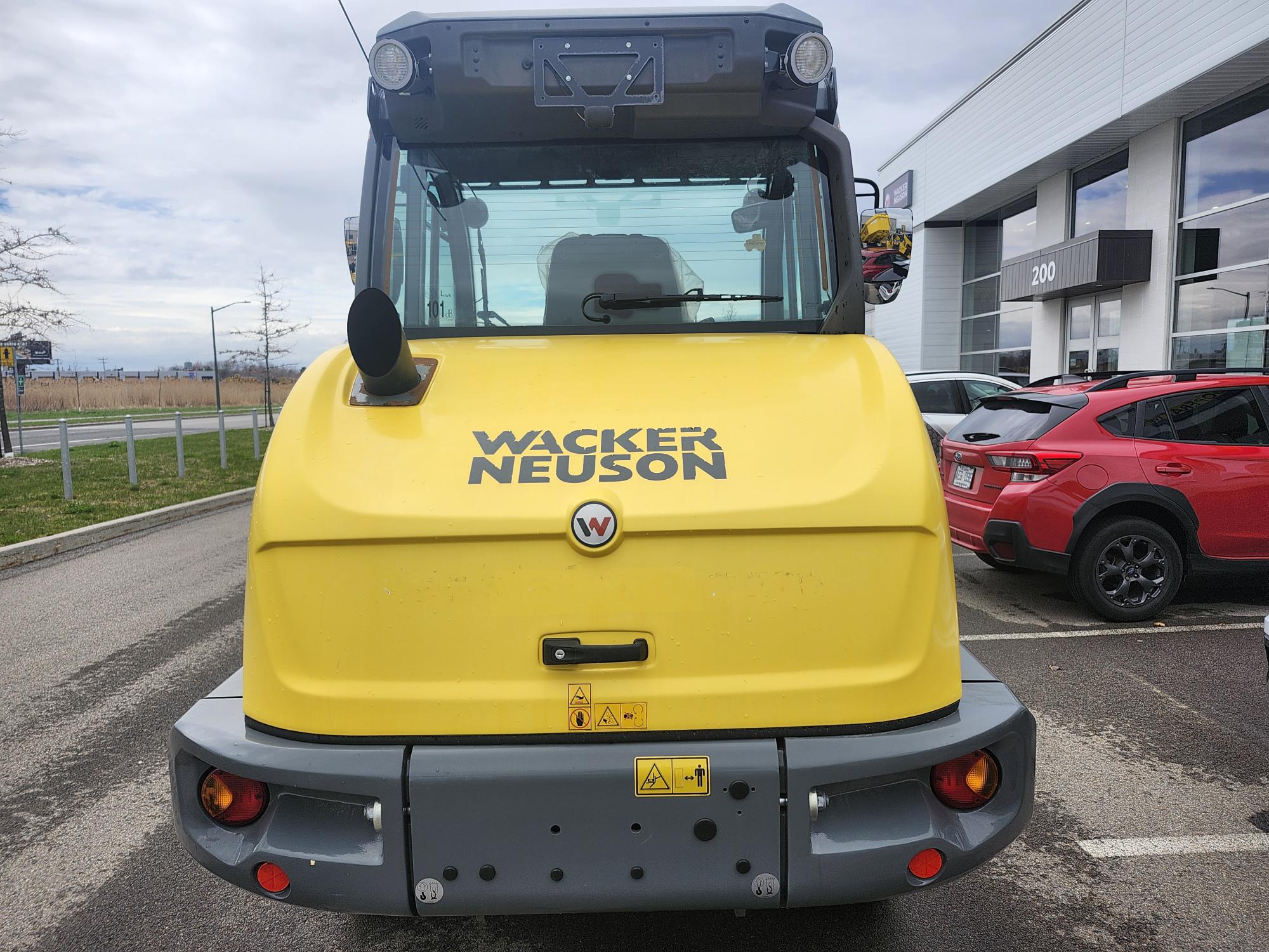Un engin de chantier jaune Wacker Neuson est garé sur un parking, près d'une voiture rouge et d'un bâtiment aux grandes fenêtres. La photo a été prise de l'arrière du véhicule.