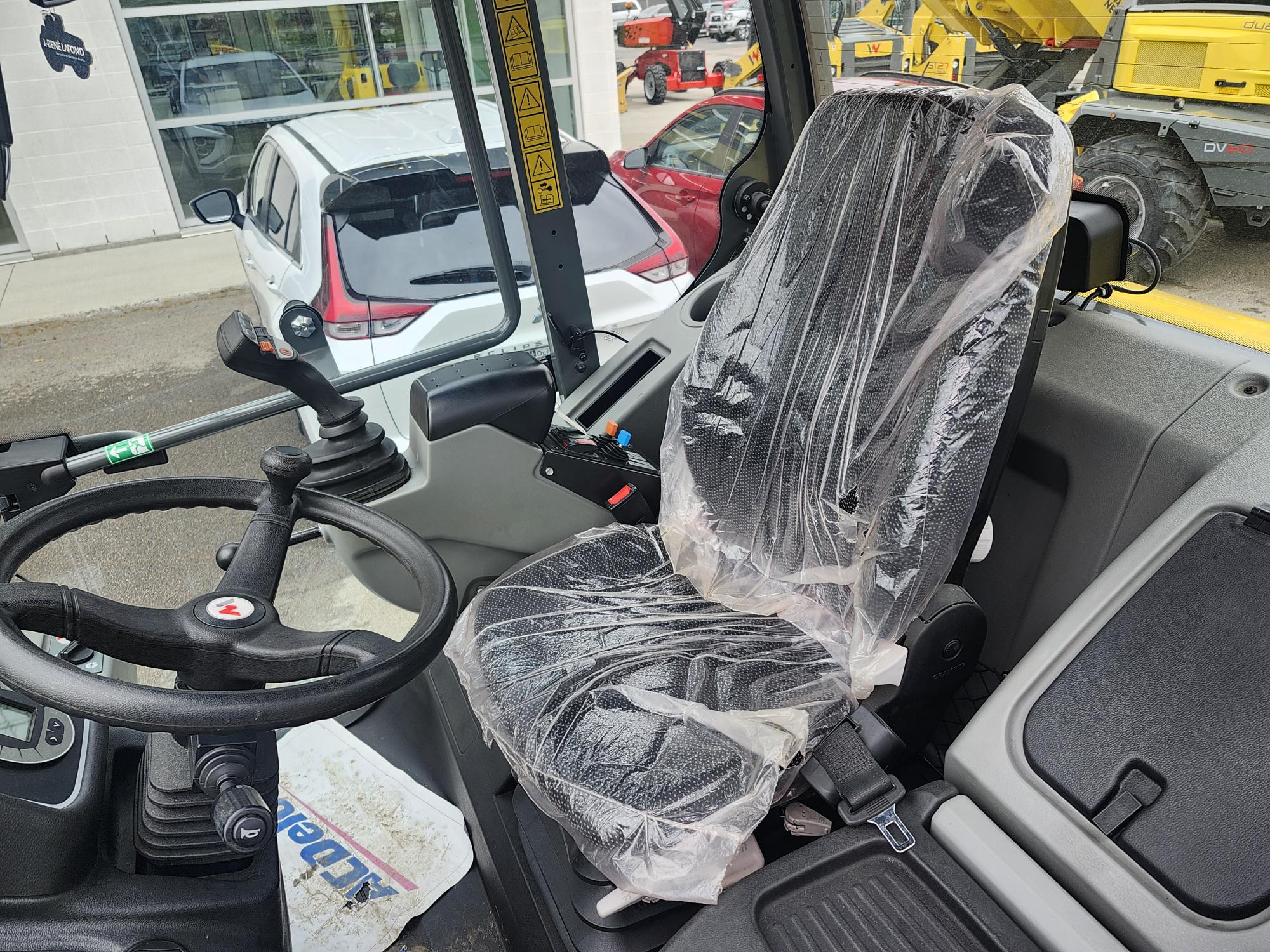 L'intérieur de la cabine d'un engin de chantier présente un siège conducteur recouvert de plastique protecteur, un volant, des leviers de commande et des pédales. D'autres véhicules et engins de chantier sont visibles à l'extérieur par les fenêtres.