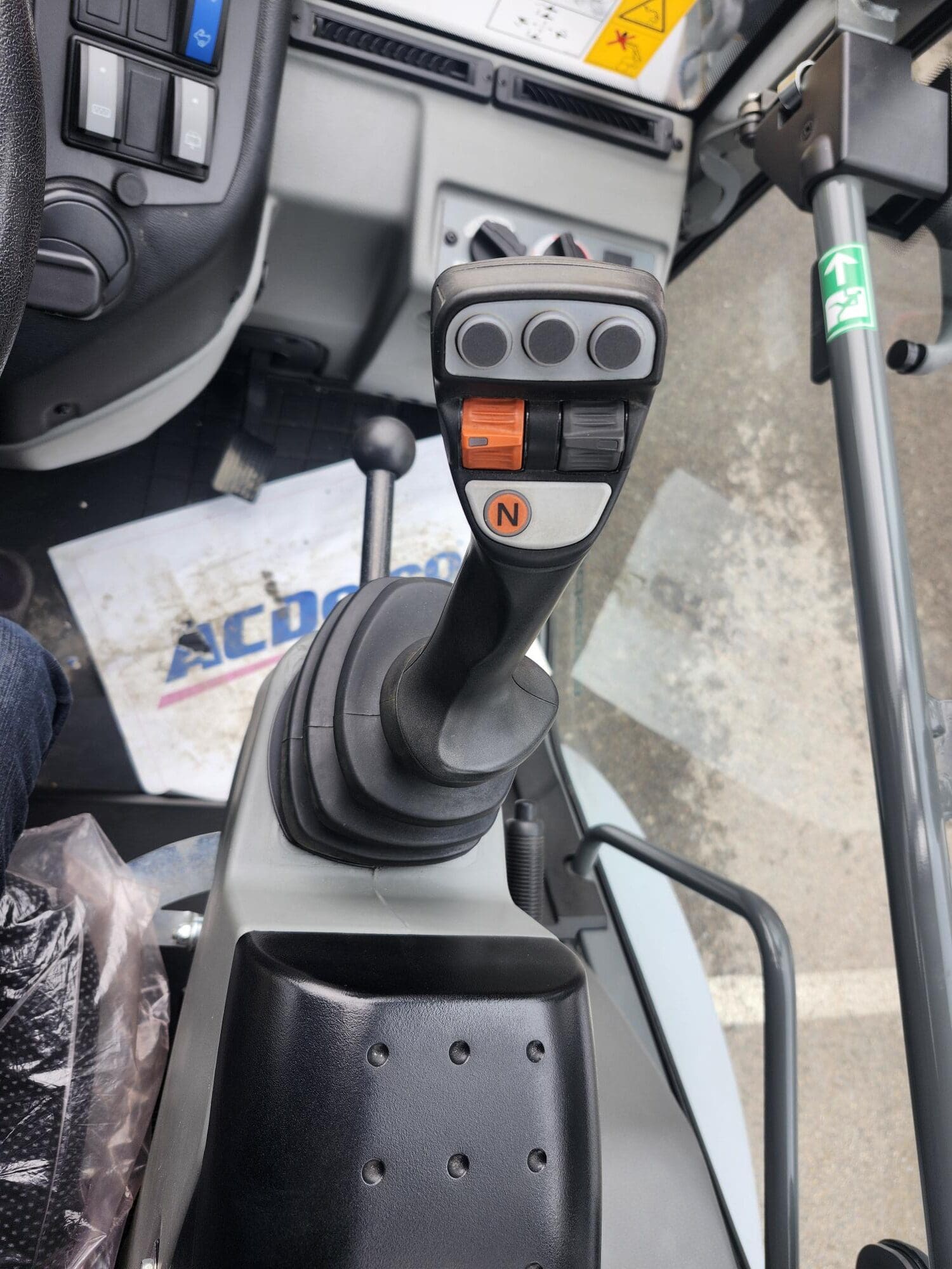 Vue rapprochée d'un joystick de commande avec plusieurs boutons et un bouton orange « N », situé à l'intérieur de la cabine d'une machinerie lourde, avec une partie du tableau de bord et du tapis de sol visibles.