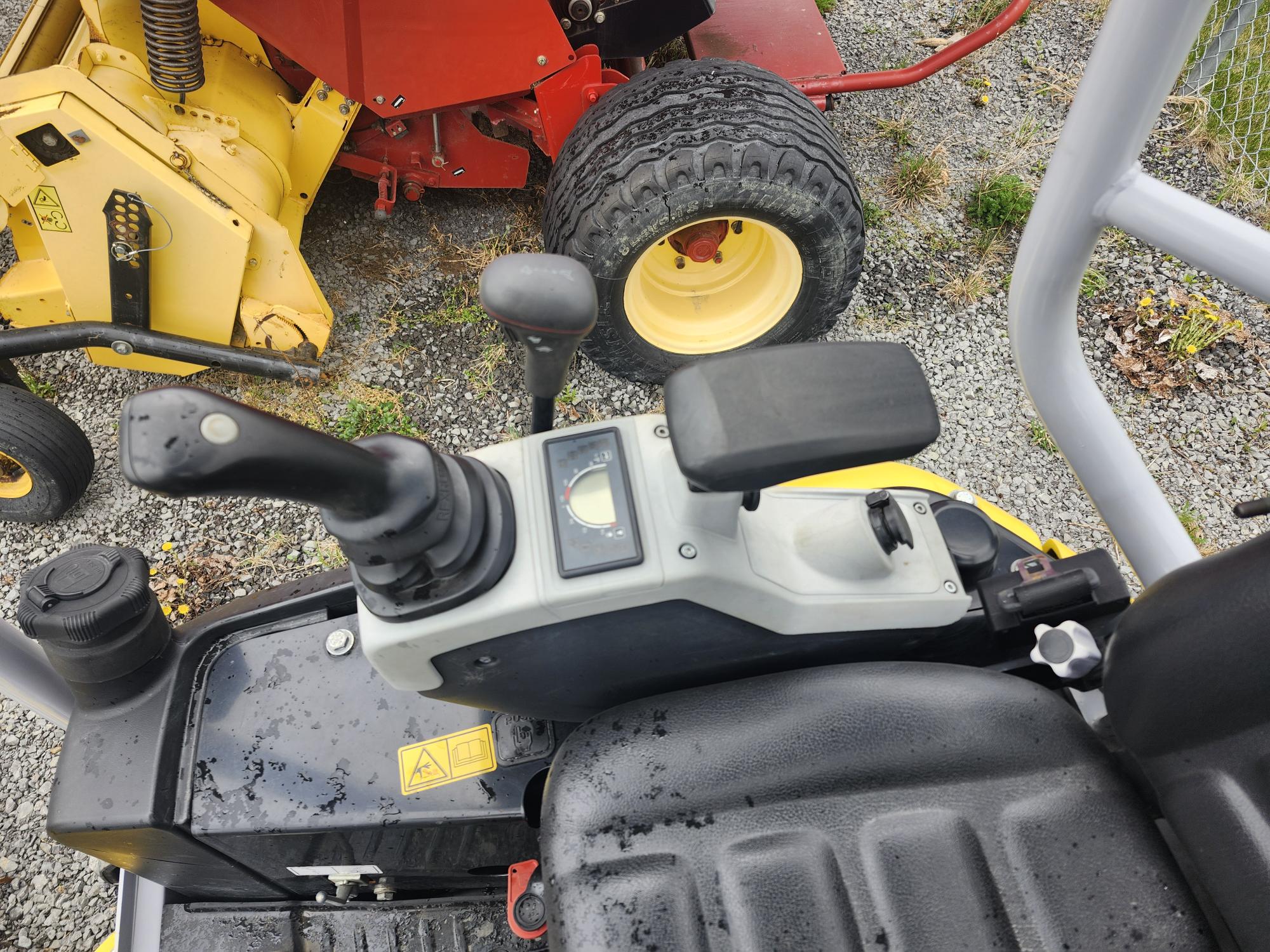 Panneau de commande et siège d'un petit engin de chantier, avec leviers, boutons et écran. Un engin plus grand, jaune et rouge, équipé d'un gros pneu, est visible en arrière-plan sur un sol en gravier.