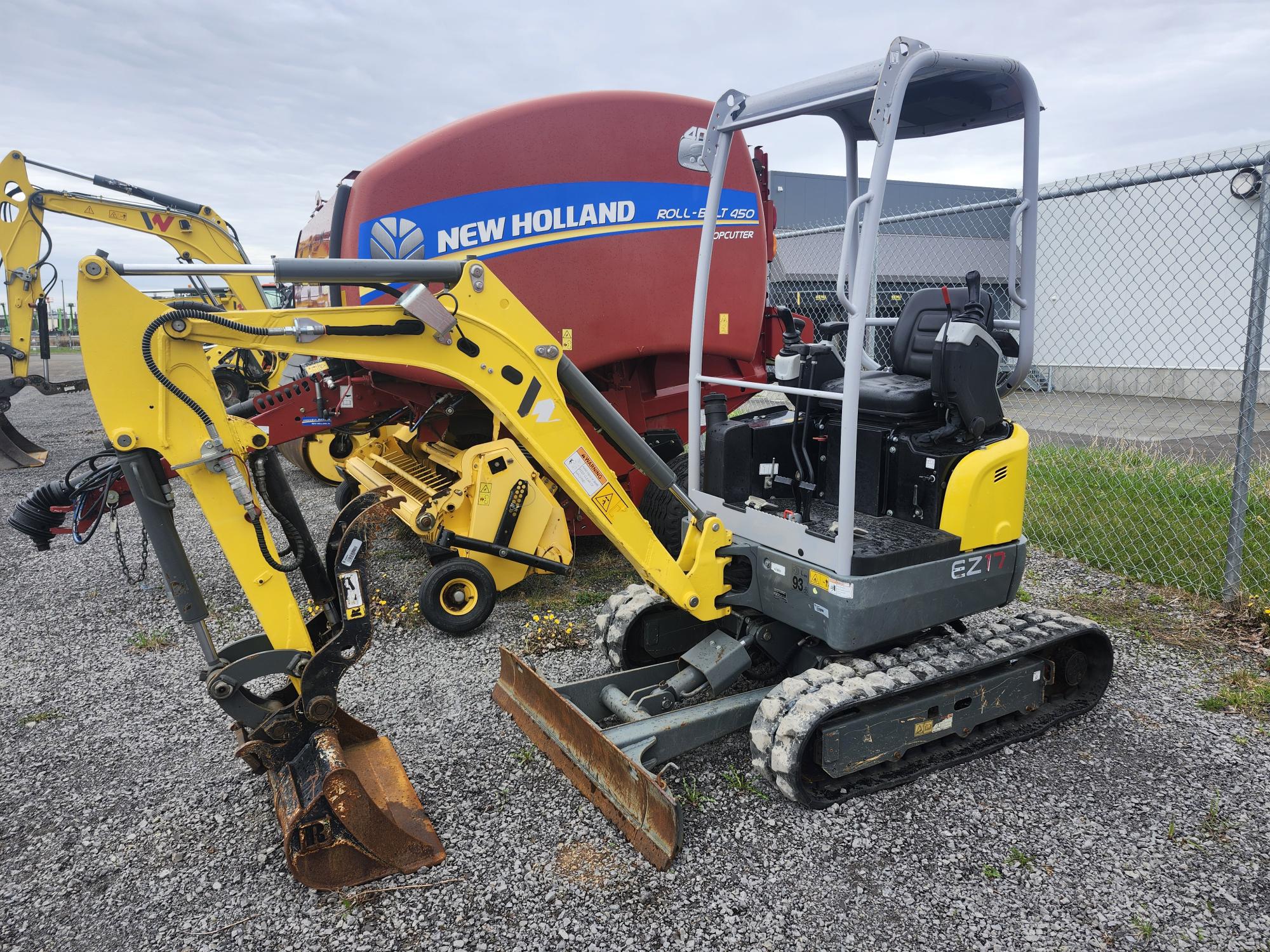 Une petite mini-pelle EZ17 jaune et noire avec un godet de creusement est garée sur du gravier à côté d'une grande machine agricole New Holland rouge près d'une clôture grillagée.
