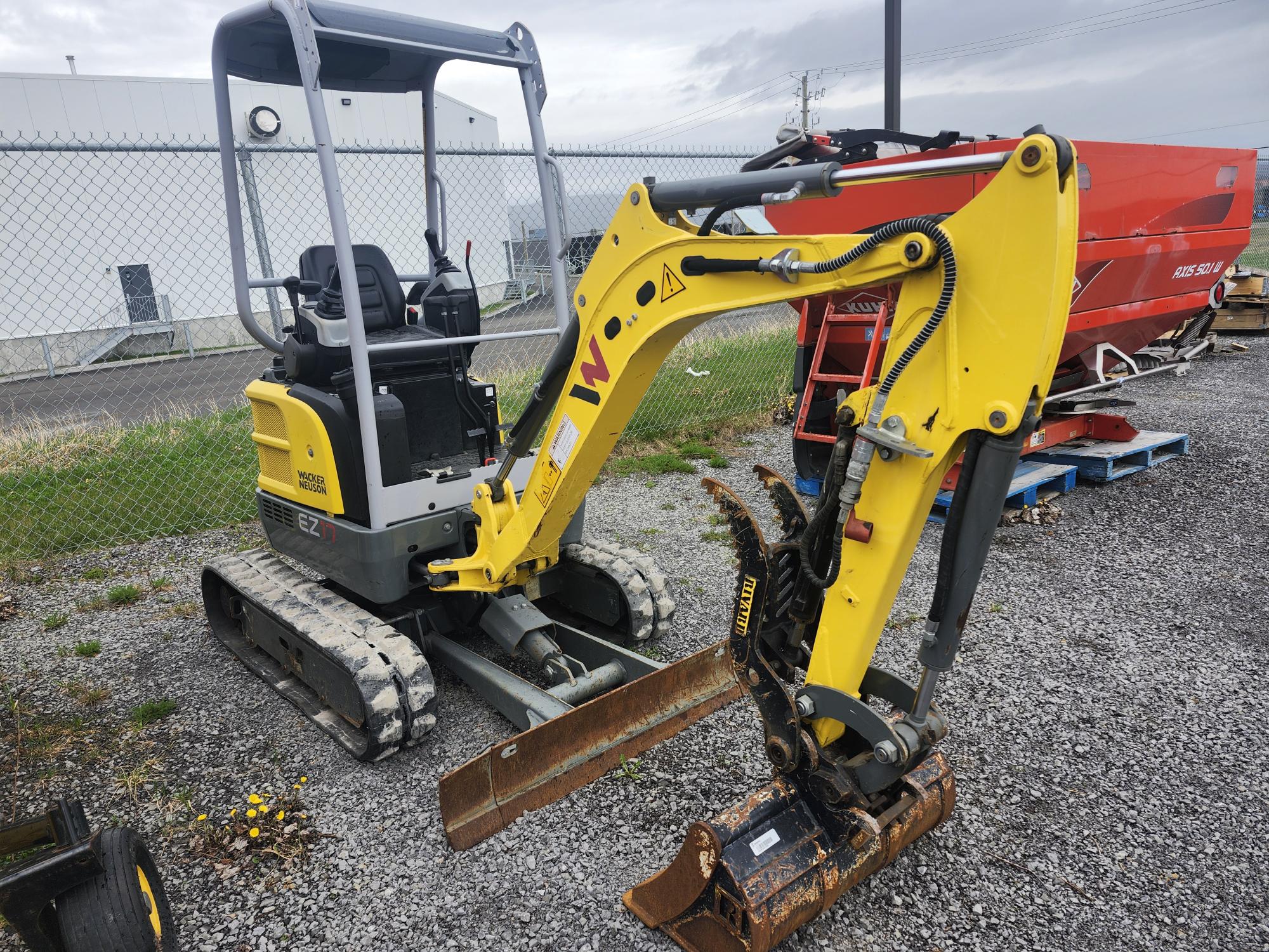 Une petite mini-pelle jaune et grise est posée sur du gravier près d'une clôture grillagée, avec une machine rouge en arrière-plan et quelques mauvaises herbes poussant à proximité.