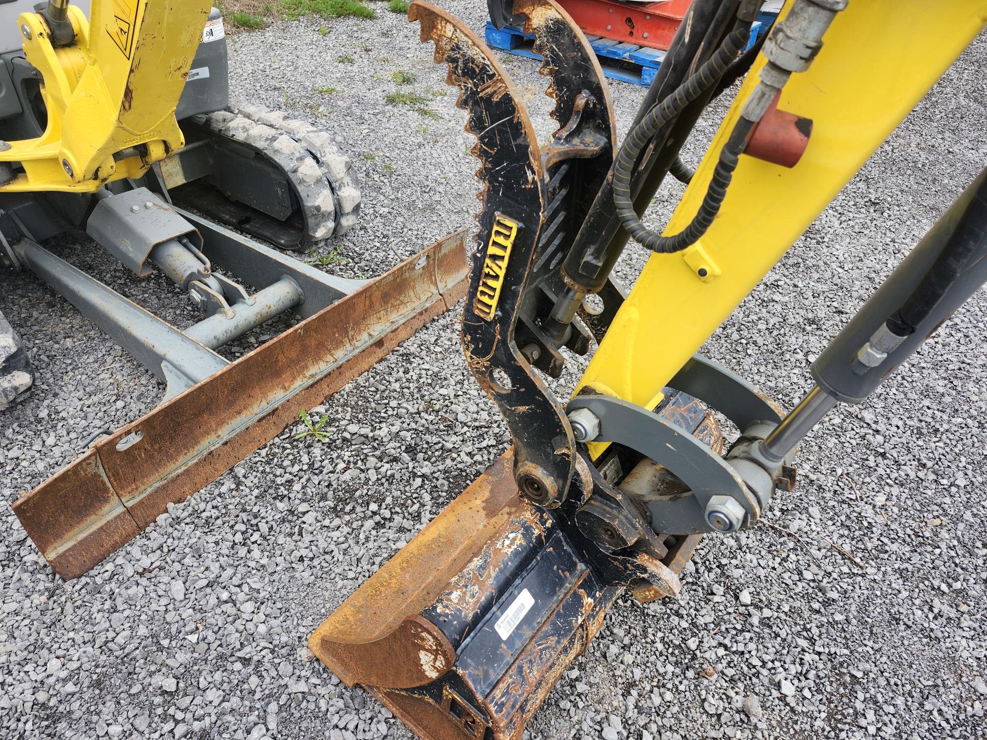 Gros plan sur l'accessoire hydraulique et le godet d'une excavatrice jaune posés sur du gravier. On aperçoit en arrière-plan une partie d'une autre excavatrice et une lame métallique. L'équipement semble usagé, avec quelques traces de rouille.