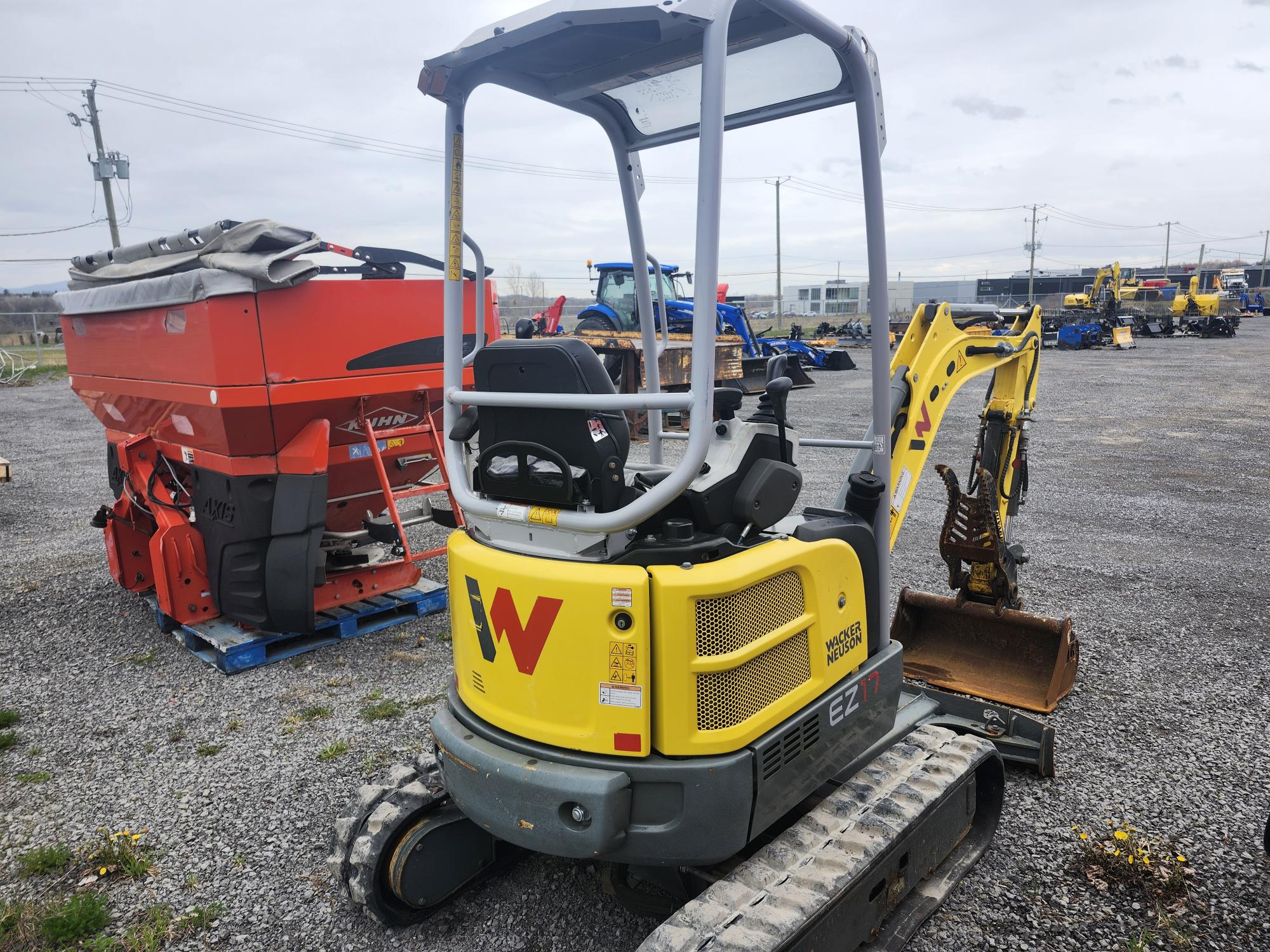 Une petite mini-pelle Wacker Neuson EZ17 jaune et grise est garée sur du gravier, près d'une machine agricole rouge et d'autres équipements, sur un terrain extérieur. Le bras et le godet de la machine sont orientés vers l'avant.
