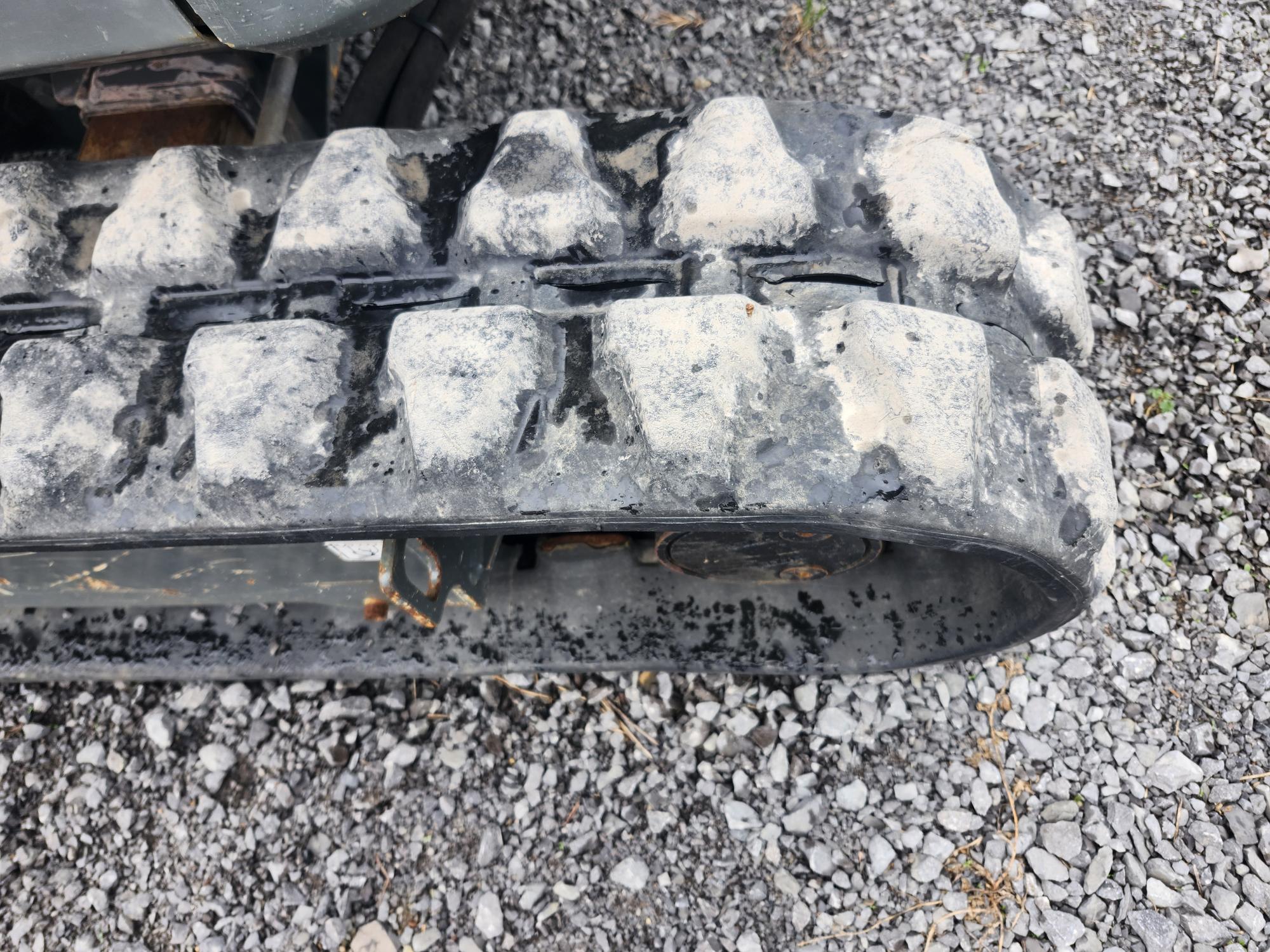 Gros plan d'une roue à chenilles en caoutchouc d'un équipement de construction reposant sur du gravier, montrant la poussière et l'usure sur la surface de la chenille.