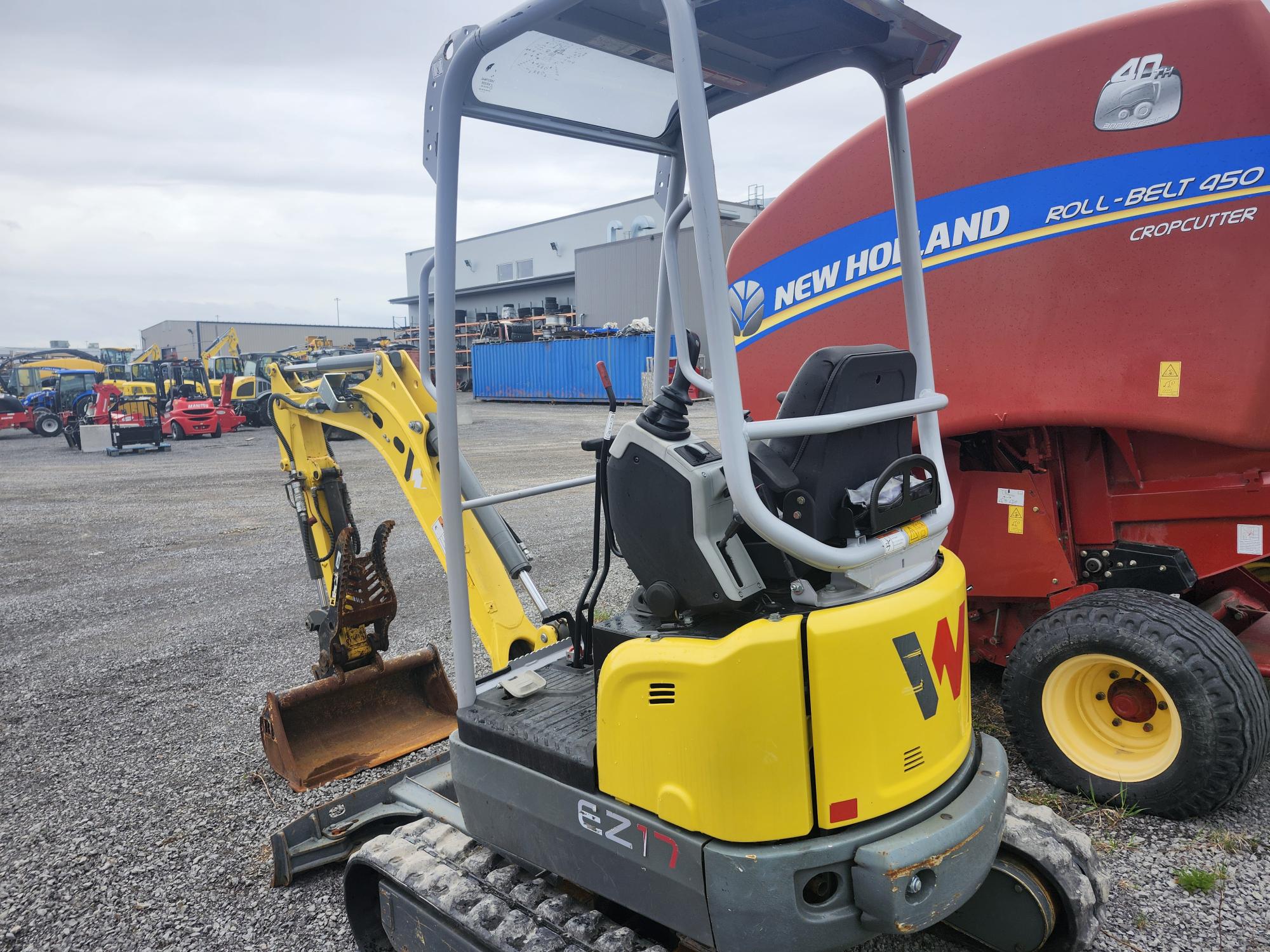 Une petite excavatrice jaune et grise est garée sur du gravier à côté d'une grande presse à balles rondes New Holland rouge, avec d'autres machines lourdes et des bâtiments industriels visibles en arrière-plan.