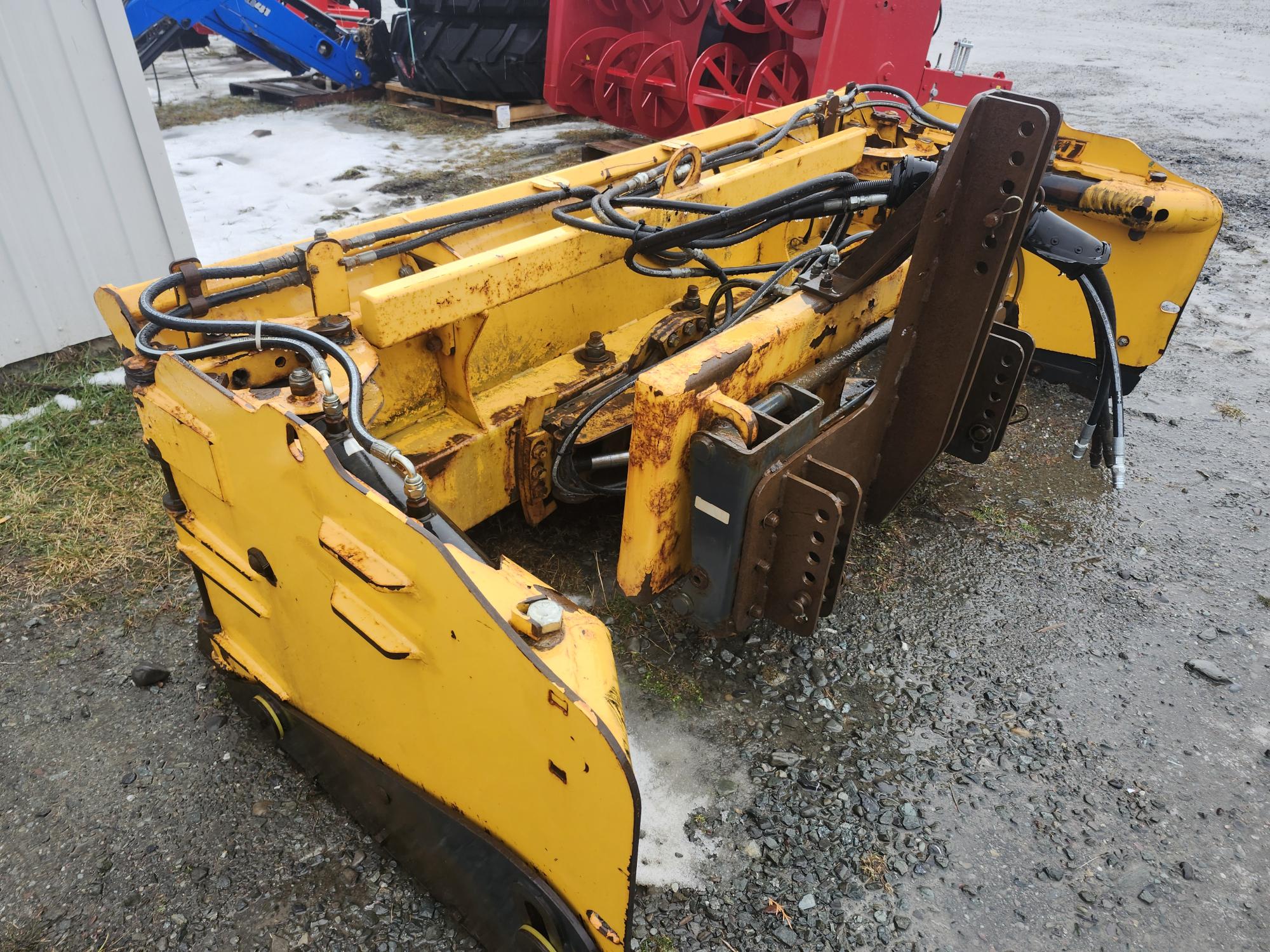 Un chasse-neige jaune rouillé équipé de composants hydrauliques est stationné sur du gravier. D'autres machines et équipements sont visibles à l'arrière-plan, certains recouverts de neige.