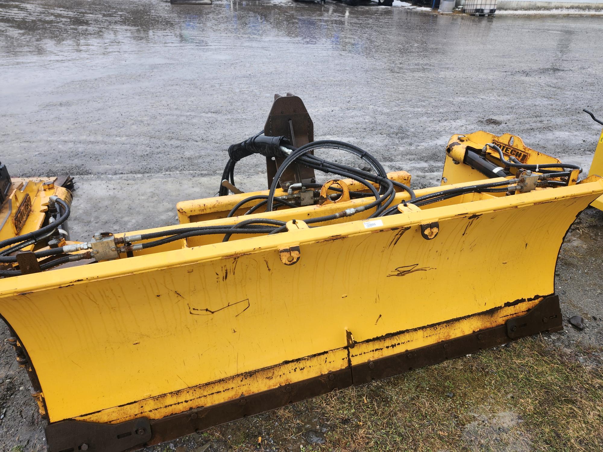 Un grand chasse-neige jaune repose sur du béton humide. Le sol est mouillé par la pluie et des tuyaux hydrauliques sont visibles sur le chasse-neige. À l'arrière-plan, la surface est couverte de taches d'eau et de reflets lumineux.