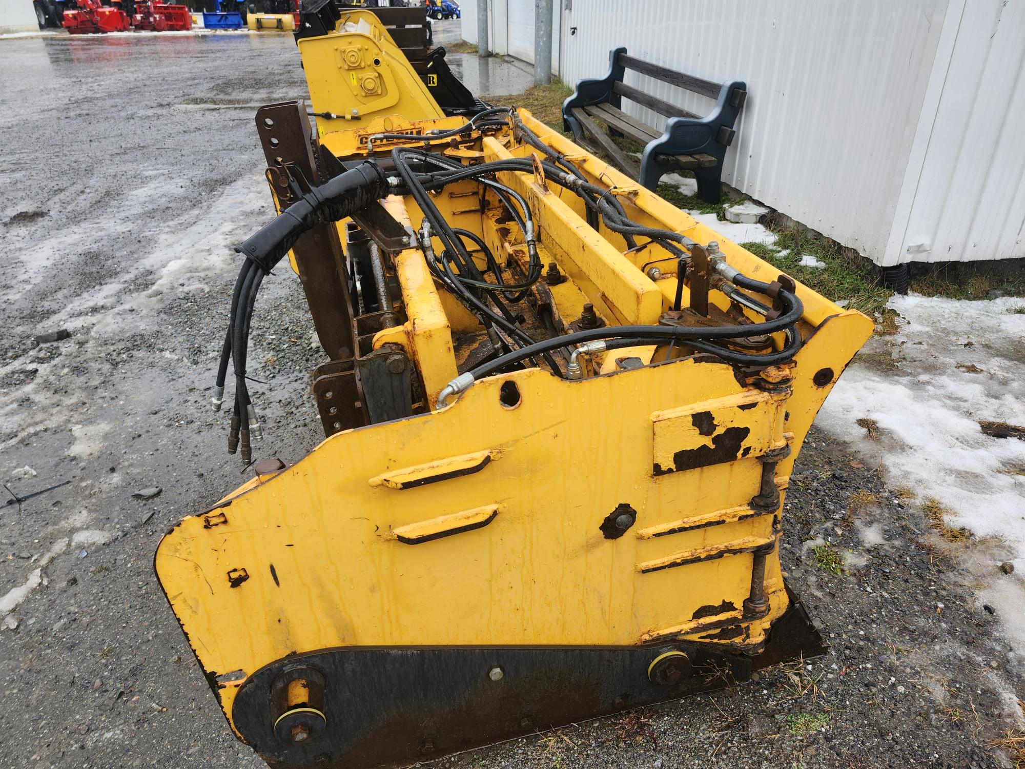 Un gros accessoire de machine industrielle jaune, peut-être un chasse-neige ou une pièce d'excavatrice, repose sur une surface humide recouverte de gravier près d'un bâtiment blanc. Un banc noir est visible à l'arrière-plan. Le métal est patiné, avec de la rouille visible et de la peinture écaillée.