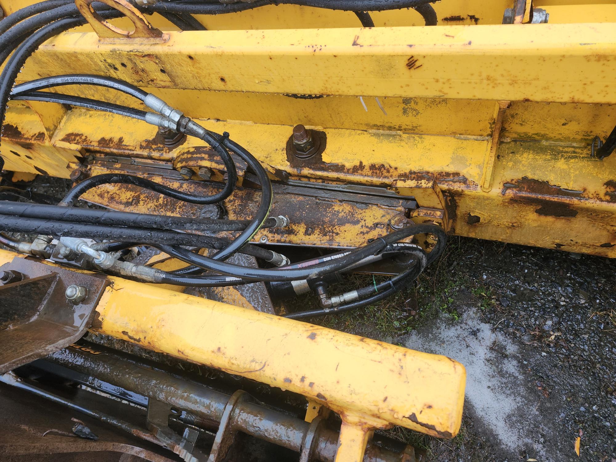 Gros plan d'une pièce de machine industrielle jaune rouillée avec des tuyaux hydrauliques noirs et des raccords métalliques. La machine est sur une surface en gravier, montrant des signes de vieillissement et d'usure.