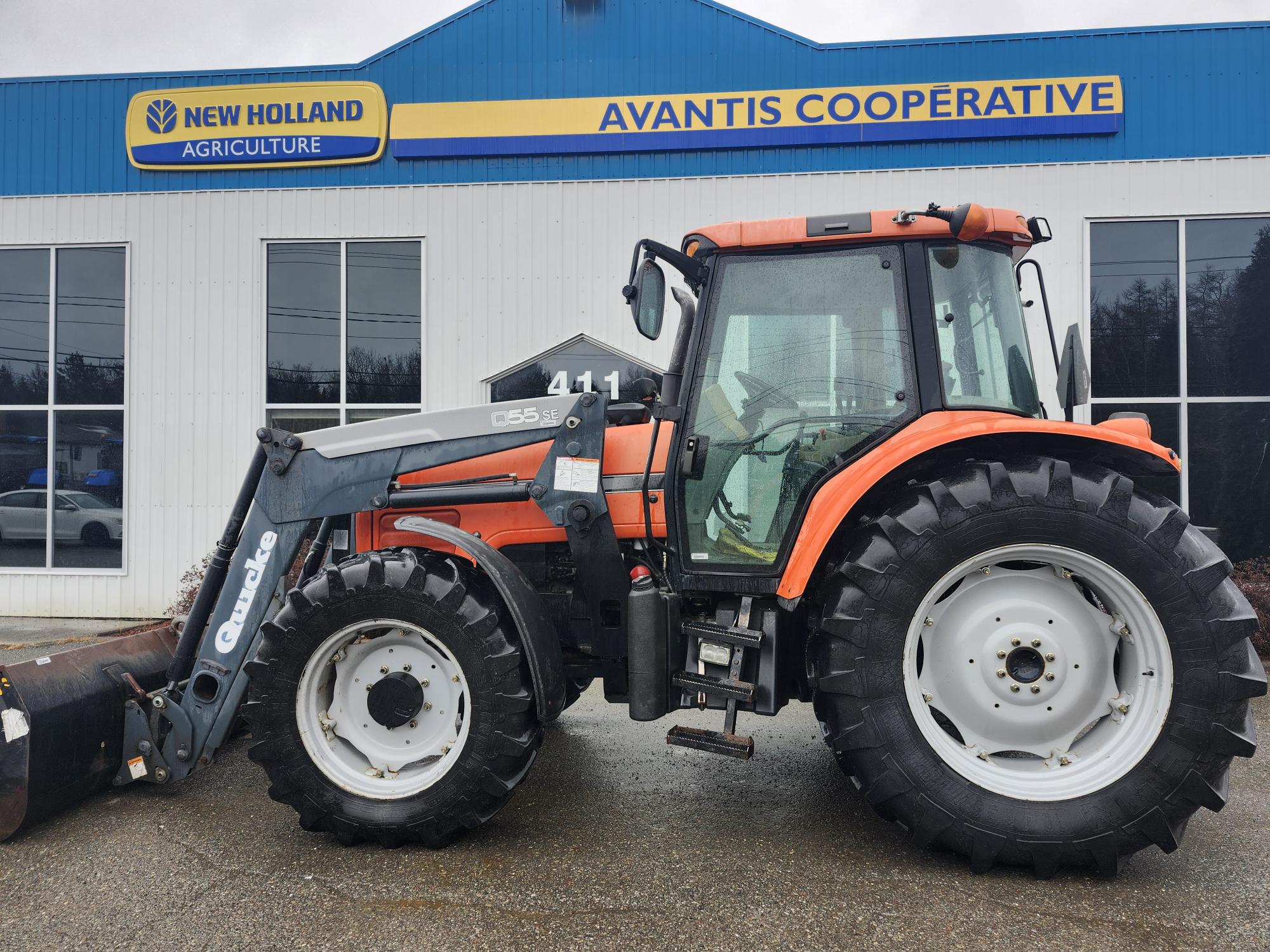 Un gros tracteur orange et noir avec de gros pneus est garé devant un concessionnaire. Le bâtiment porte un panneau bleu et jaune indiquant « New Holland Agriculture » et « Avantis Cooperative ». Le chargeur frontal du tracteur est légèrement surélevé.