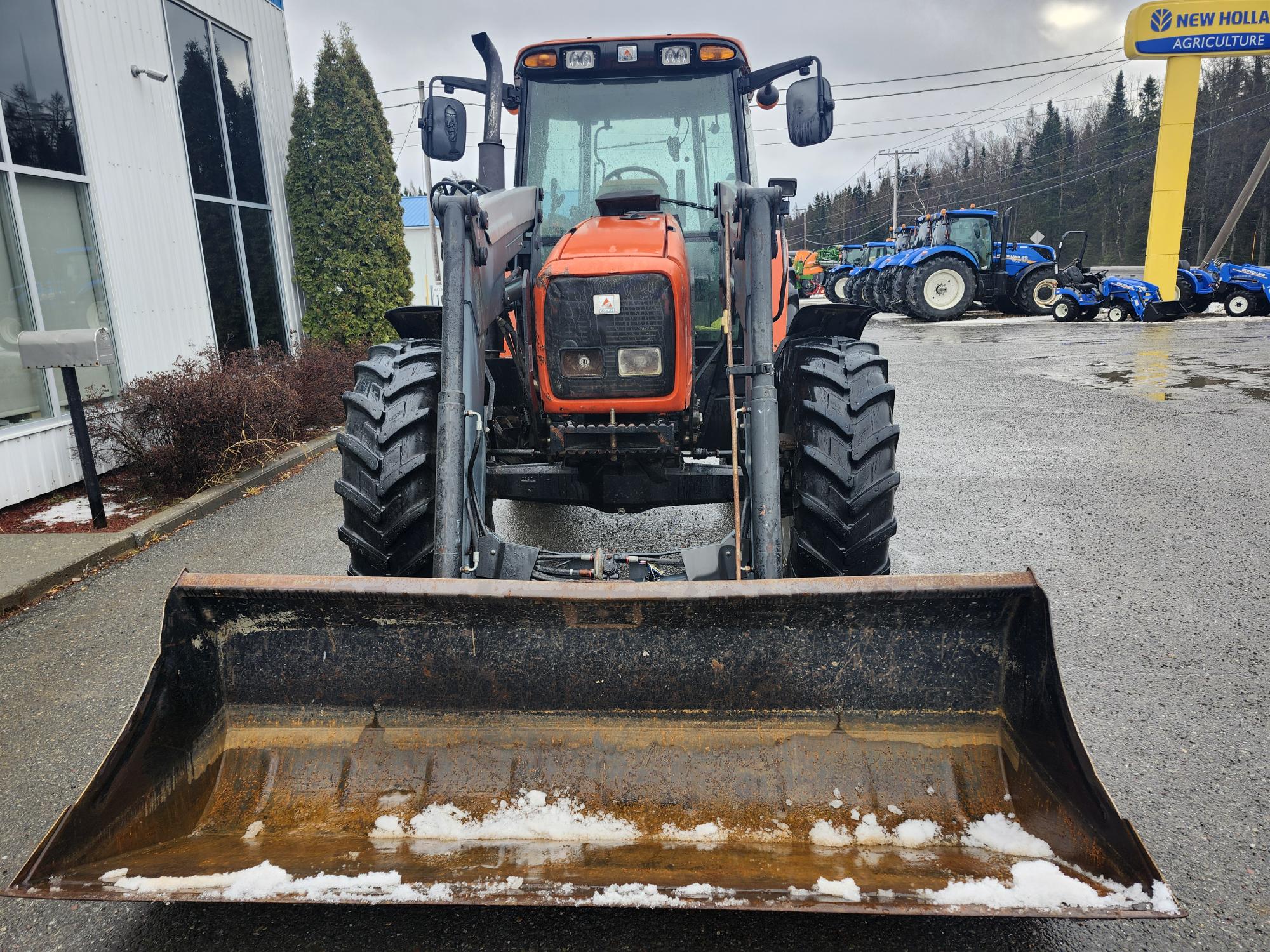 Un tracteur rouge équipé d'un chargeur frontal et de plaques de neige est stationné par une journée pluvieuse et nuageuse à l'extérieur d'un concessionnaire agricole New Holland. Plusieurs autres tracteurs sont visibles à l'arrière-plan.